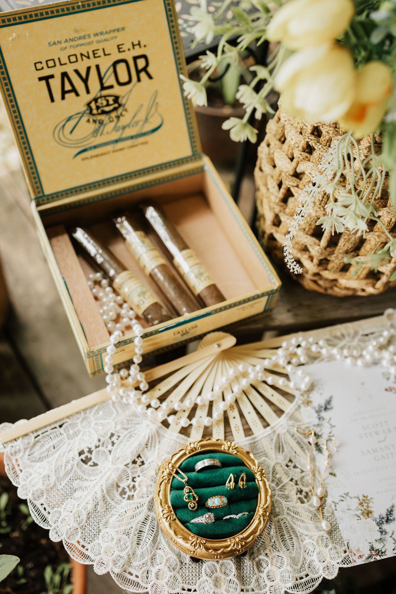 Vintage Colonel E.H. Taylor cigar box with pearls, gold fan, green velvet ring dish with jewelry, and wicker vase on lace.
