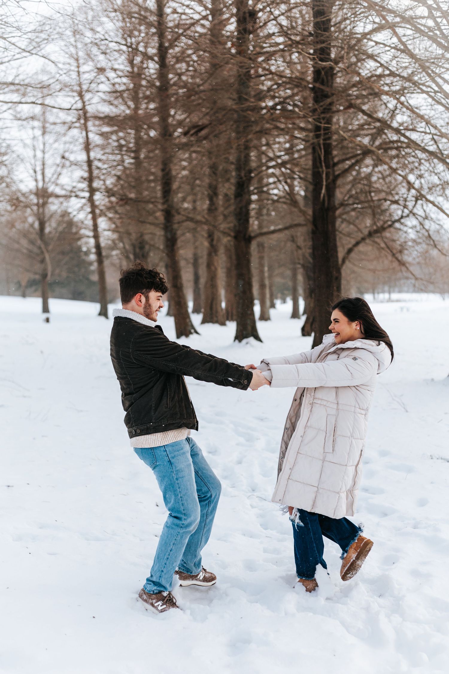 A playful couple dancing and holding hands in the snow surrounded by bare trees.