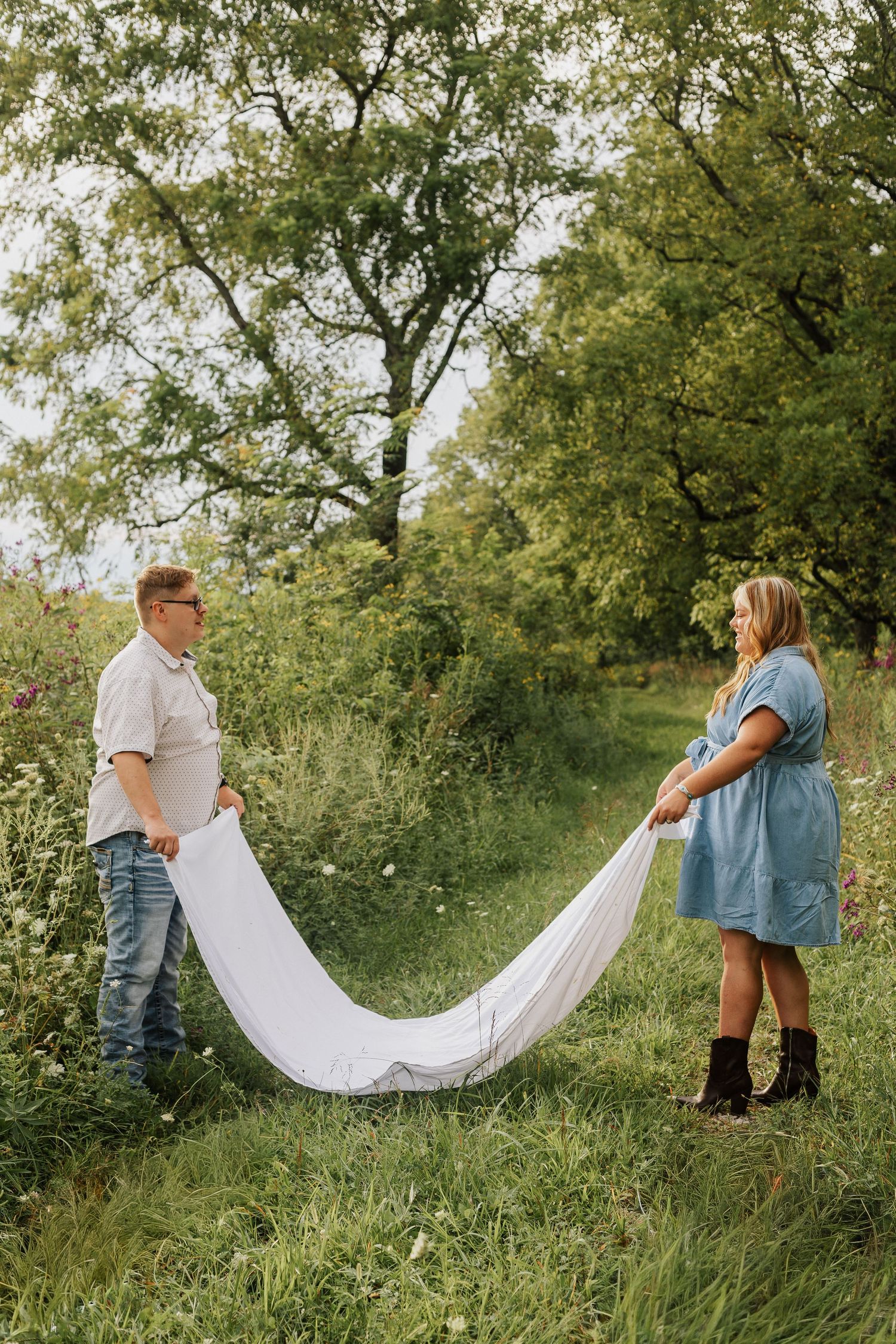 Couple holding white fabric in grassy meadow during outdoor photoshoot.