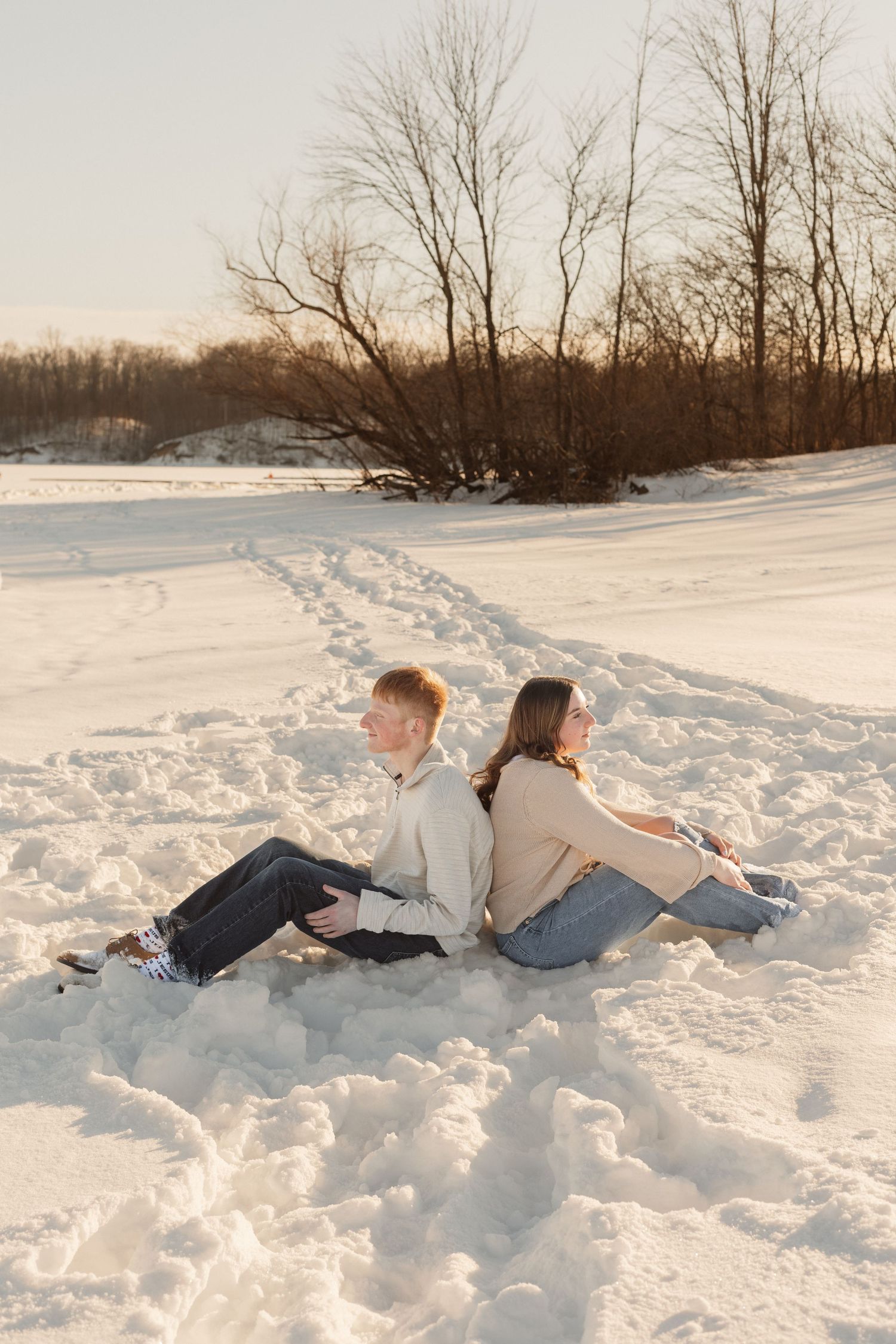 A couple sits back-to-back in the snow during a winter sunset photoshoot.
