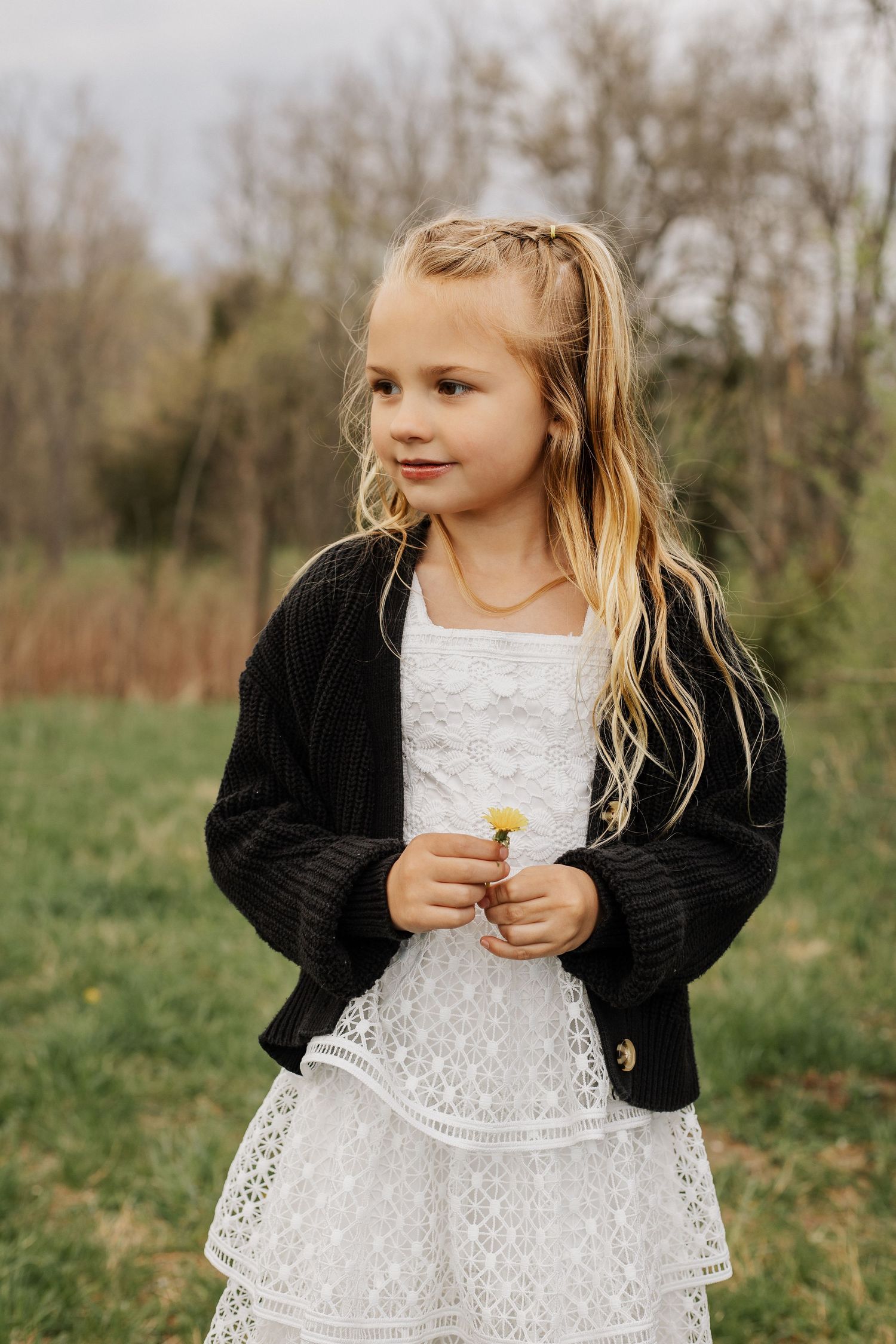 Young girl in white lace dress and black cardigan holds small yellow flower outdoors.