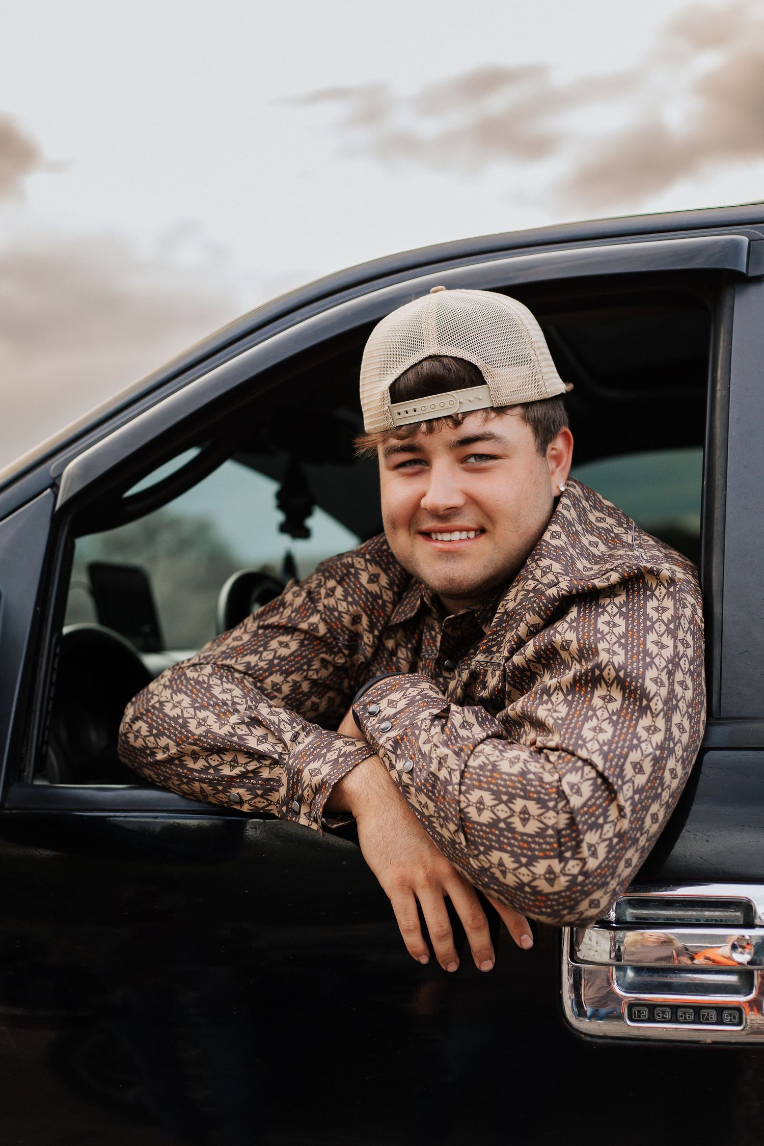 Person wearing baseball cap and patterned hoodie leans out of black truck window.