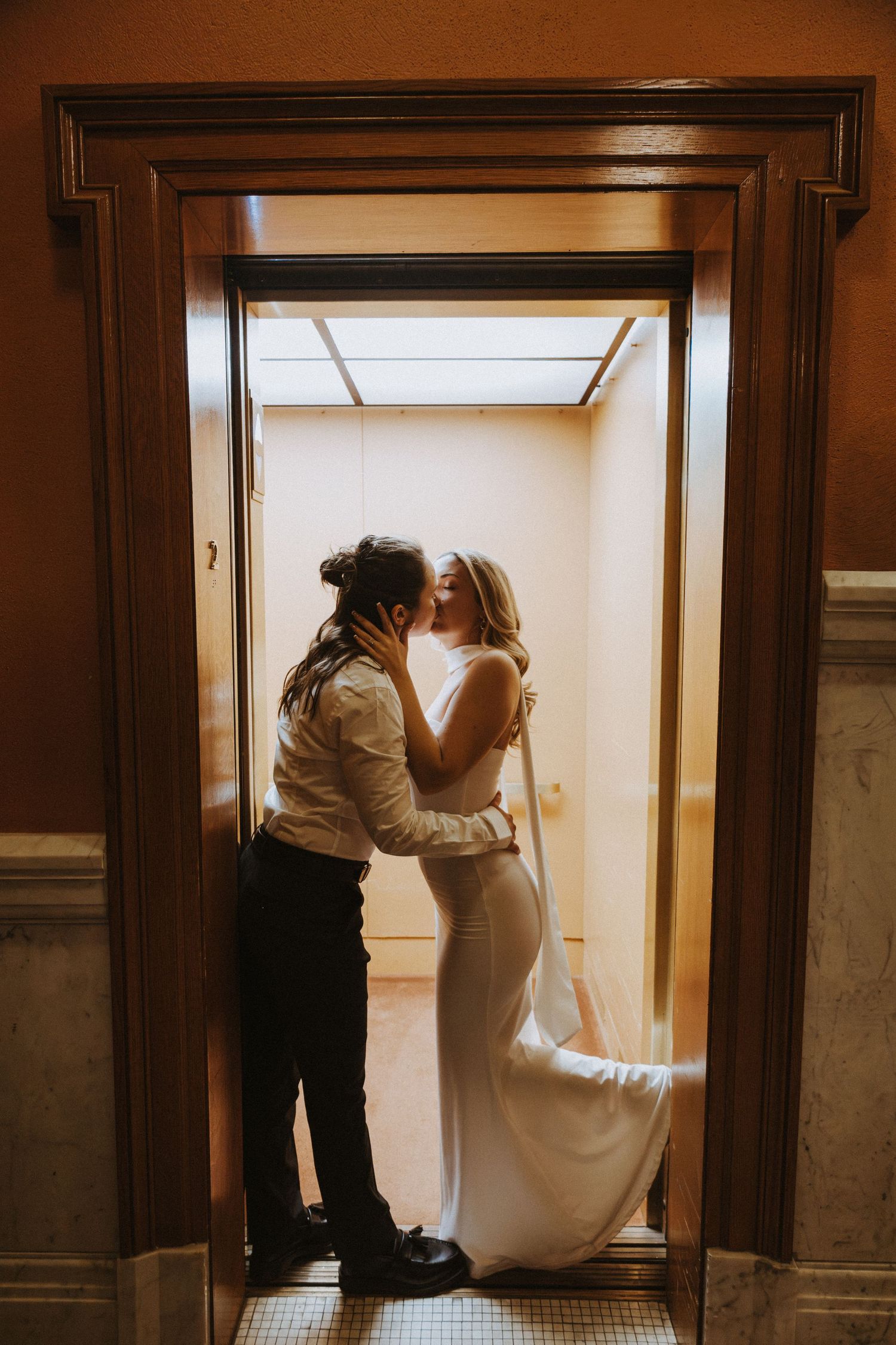 Couple sharing a romantic kiss in an elevator doorway, bride in white gown with veil, groom in dark pants and white shirt.