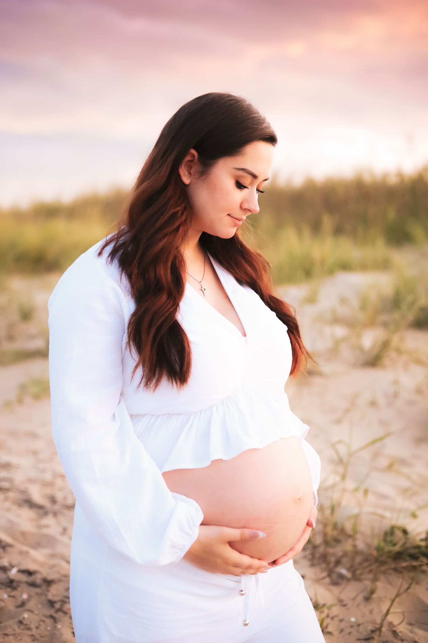 Maternity beach photoshoot with woman in white dress against pink sunset sky.