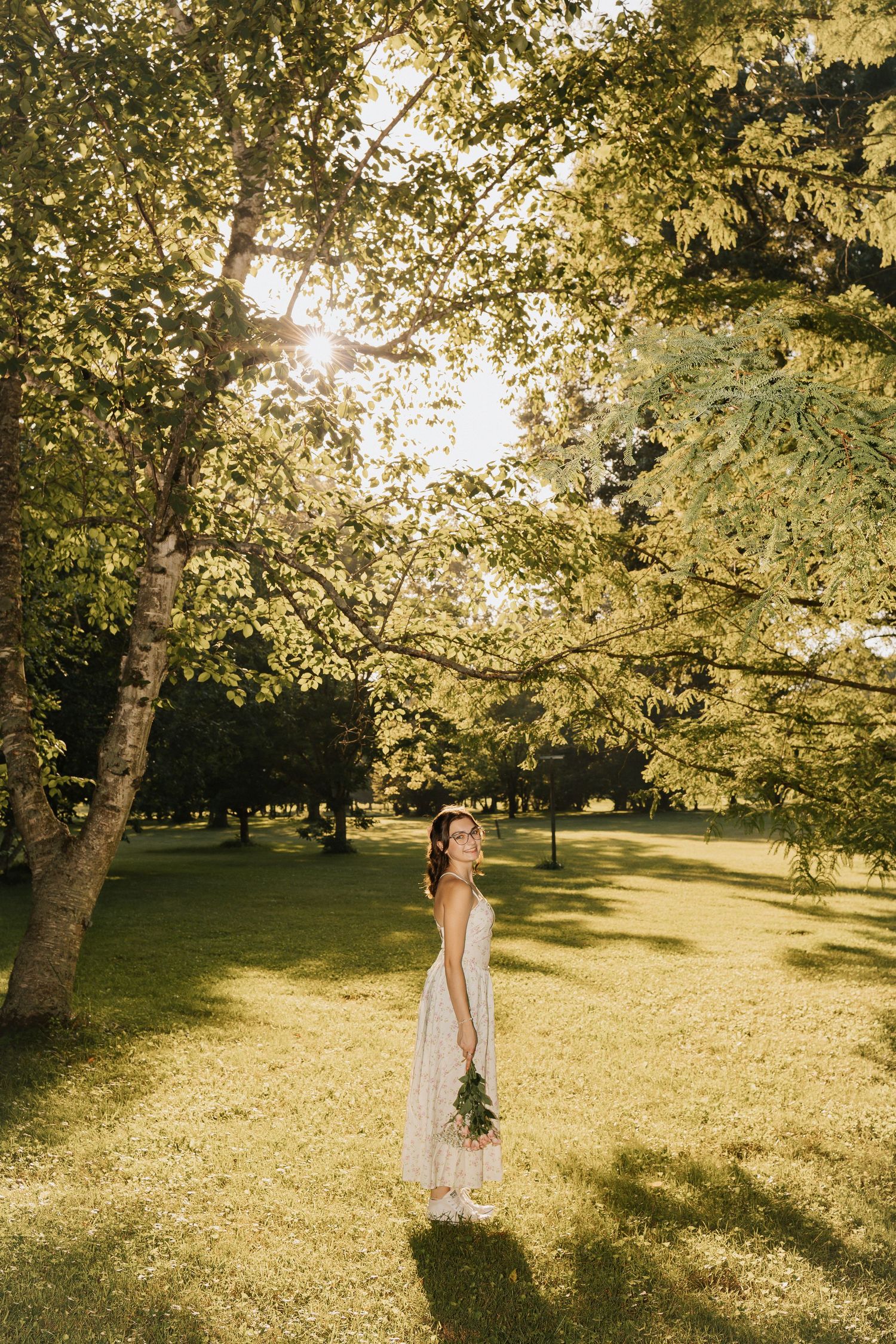 Bride in white dress holding bouquet stands in sunlit park surrounded by lush green trees on a beautiful summer day.