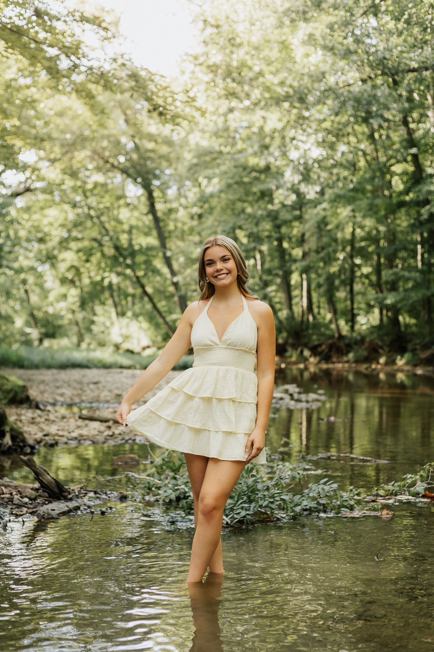 Woman in white dress playing in shallow creek water surrounded by green forest.