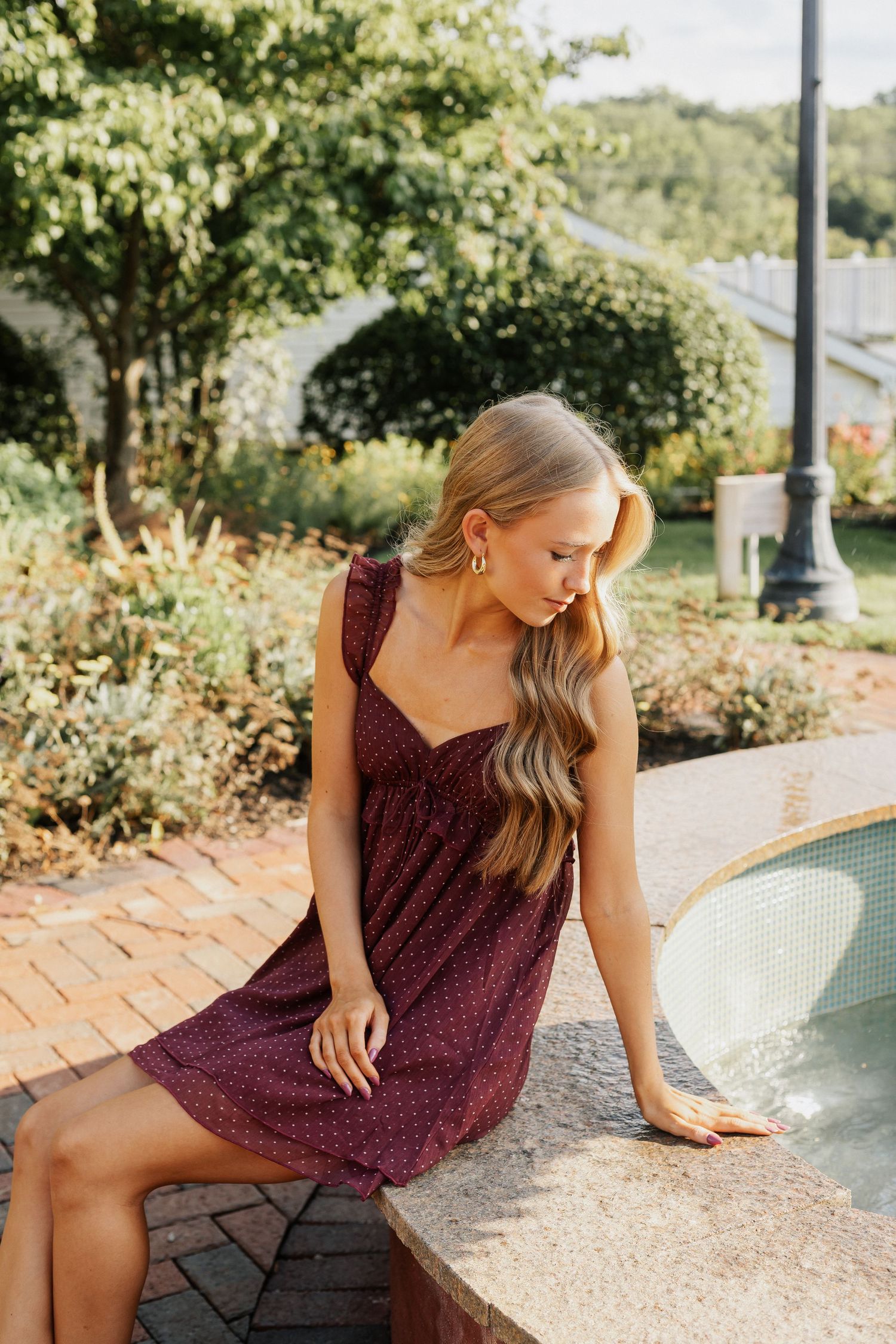 Woman in burgundy dress poses near water feature during outdoor photoshoot.