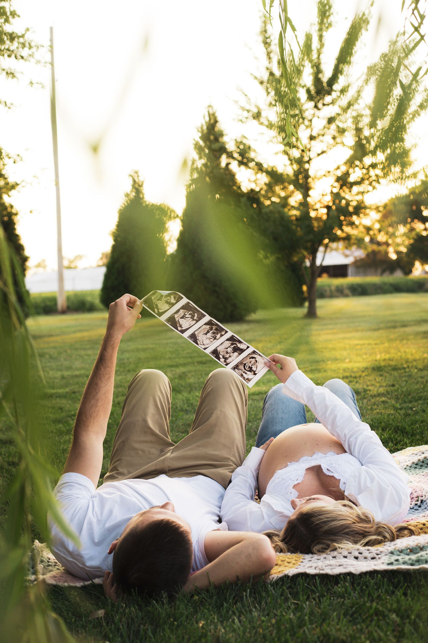 Couple lying on blanket in grass viewing ultrasound photos during sunset.