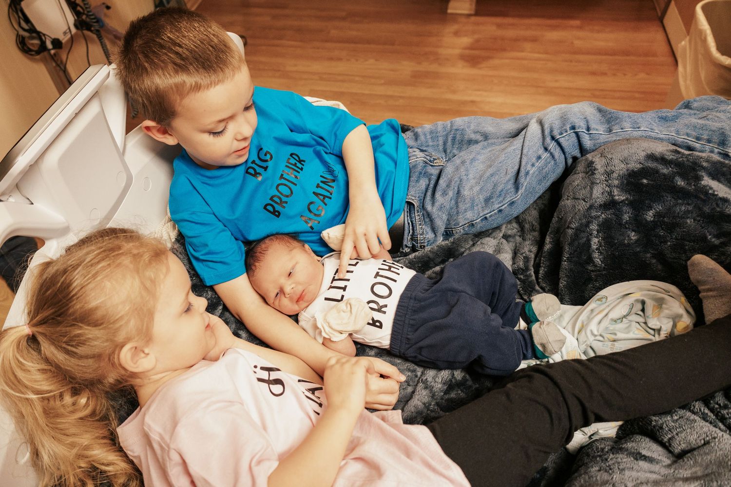 Siblings interacting playfully on hospital bed during newborn visit.