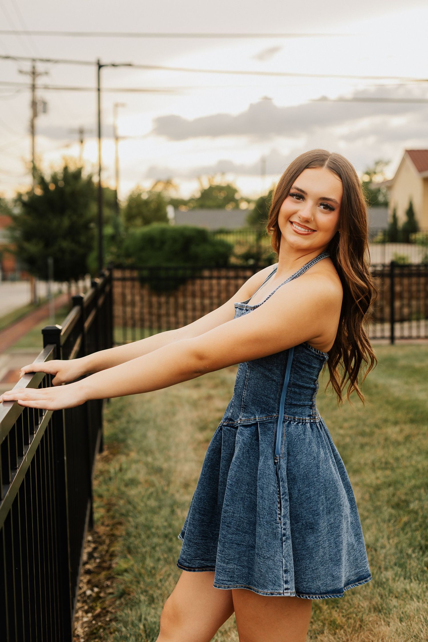 Young woman in denim dress poses by black metal railing at sunset in suburban setting.