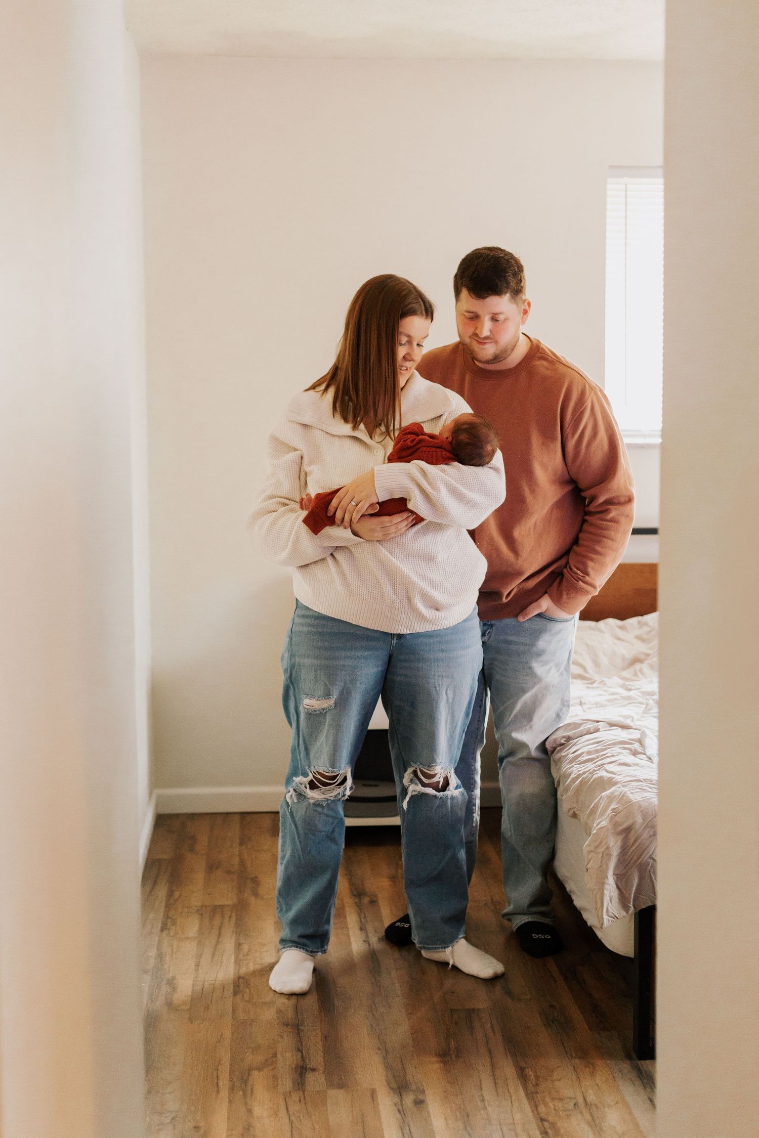 A couple embraces in a casual indoor setting with wooden floors and white walls.