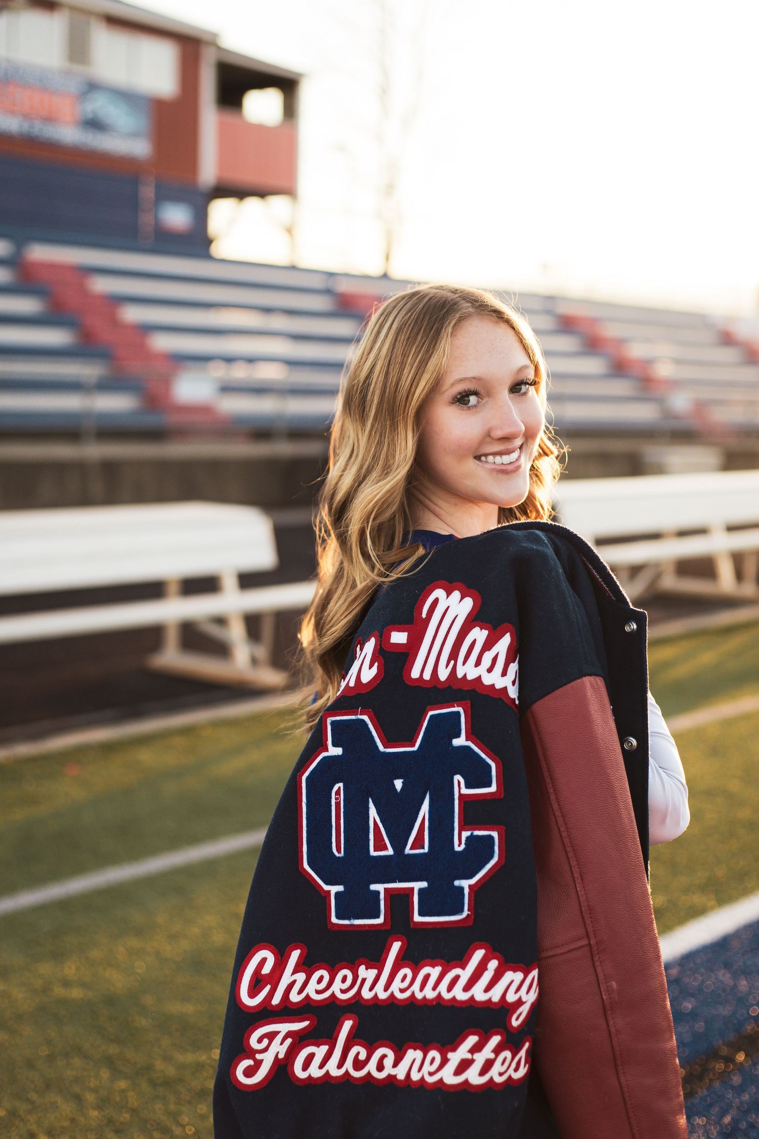 Cheerleader wearing a navy and white varsity jacket with red lettering on the back poses on a football field.