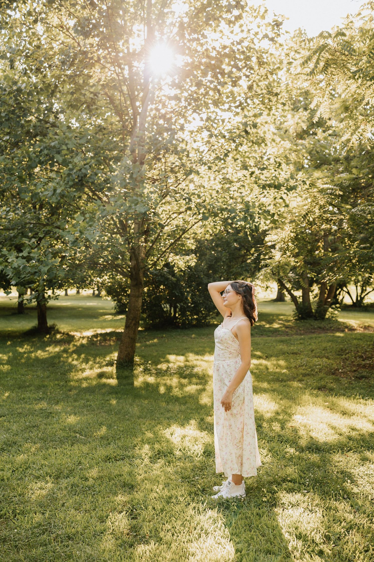 Woman in floral sundress standing in sunlit green park, looking up with hand in hair, wearing white sneakers.