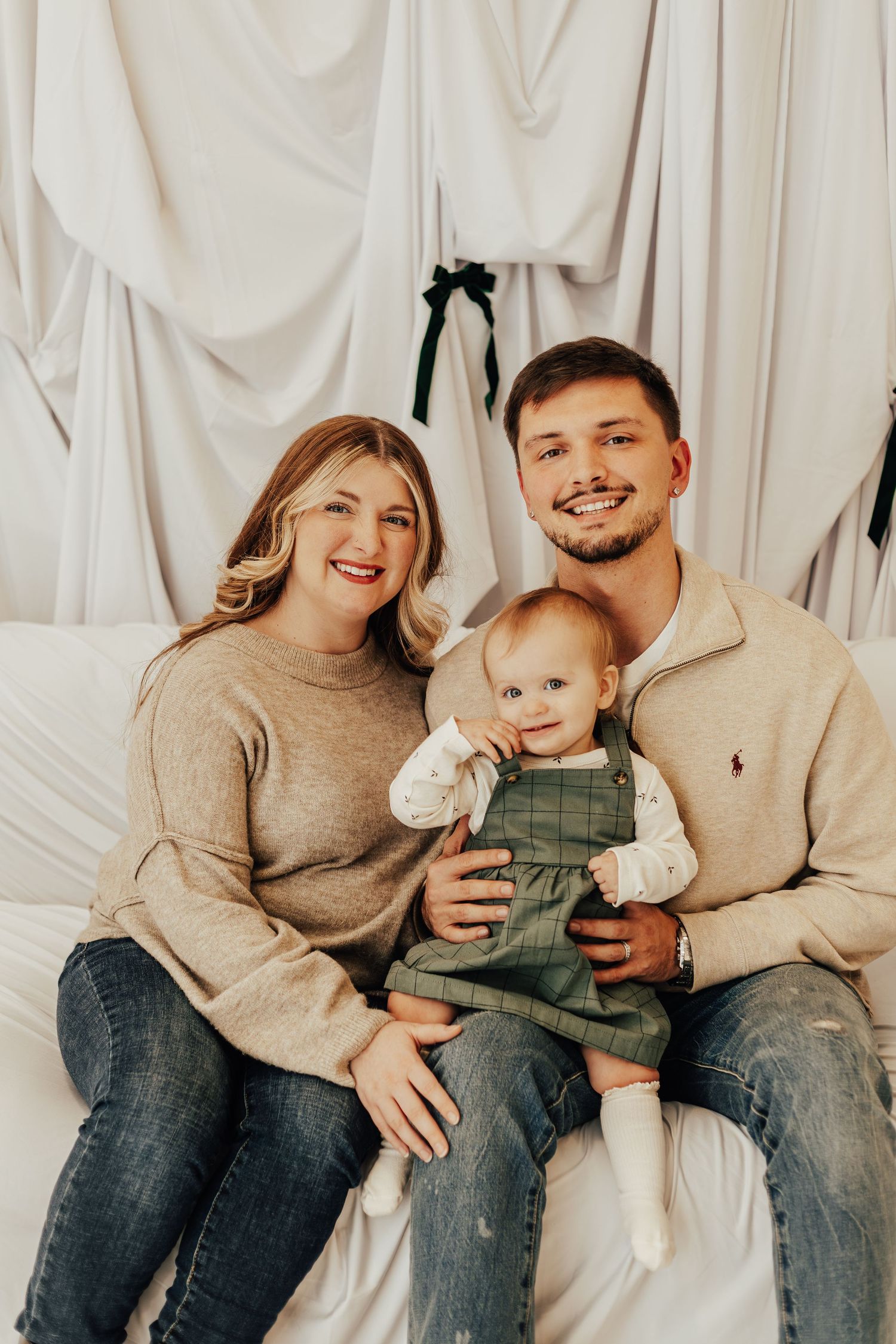 Studio family portrait with baby in overalls against white fabric backdrop.