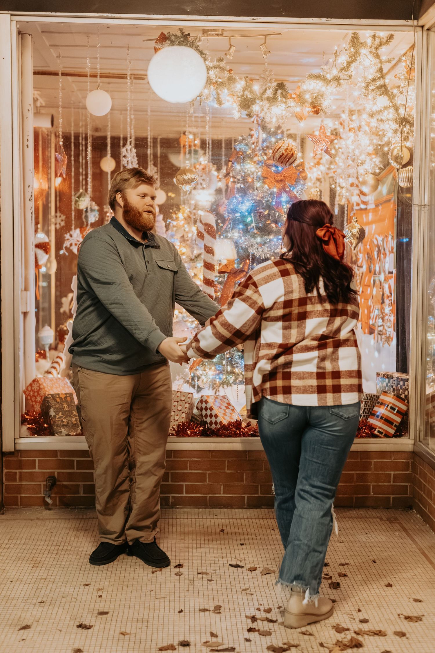 Couple in casual attire playfully dancing together against a brick wall with twinkling lights in the background.
