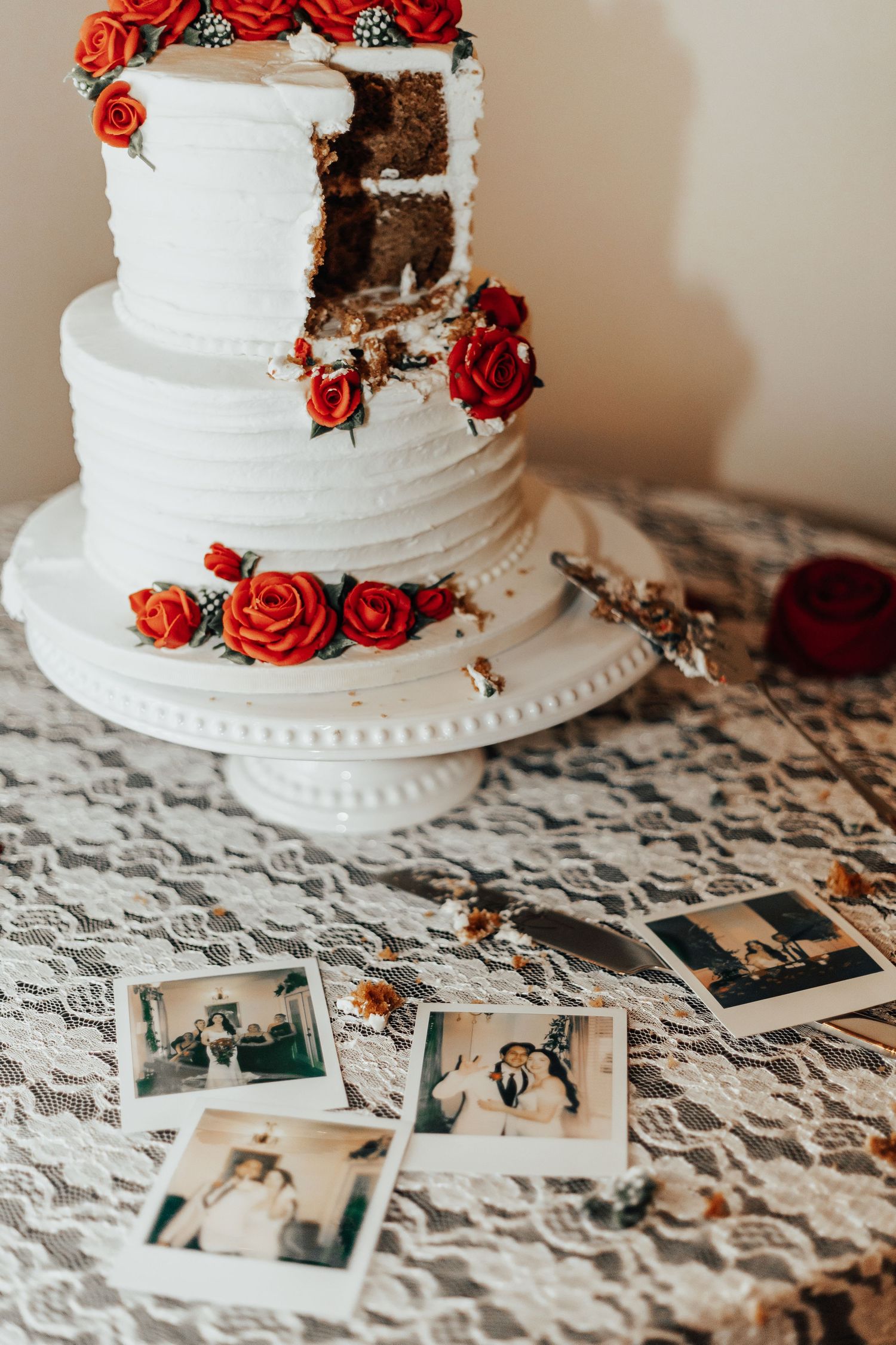 White wedding cake decorated with red roses sits on a lace tablecloth with vintage photos displayed beneath.