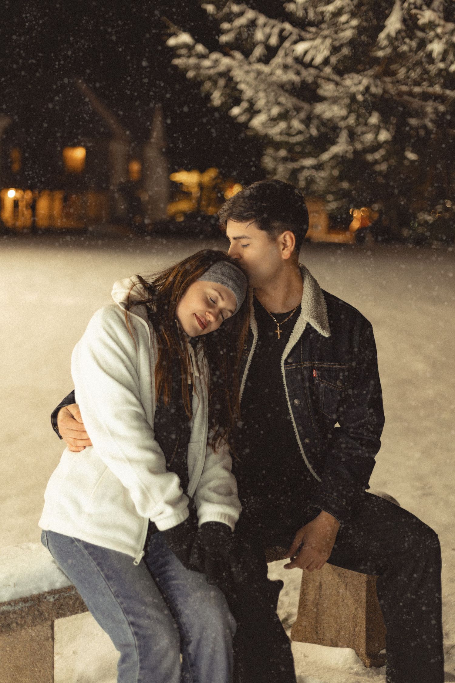 Two people sitting close together on a bench during a snowy evening.