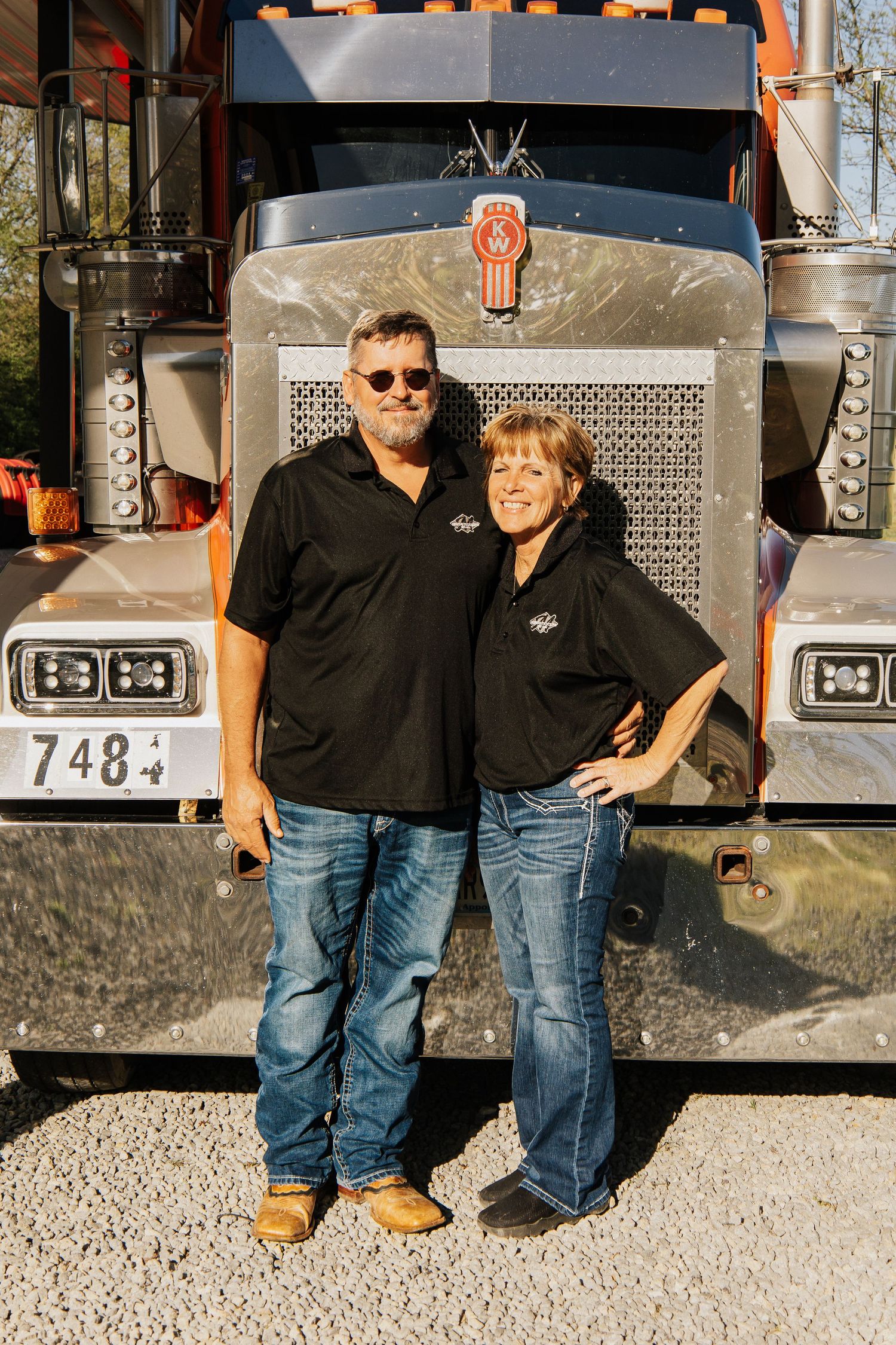 Two people in matching black polo shirts stand smiling in front of a Kenworth semi truck.