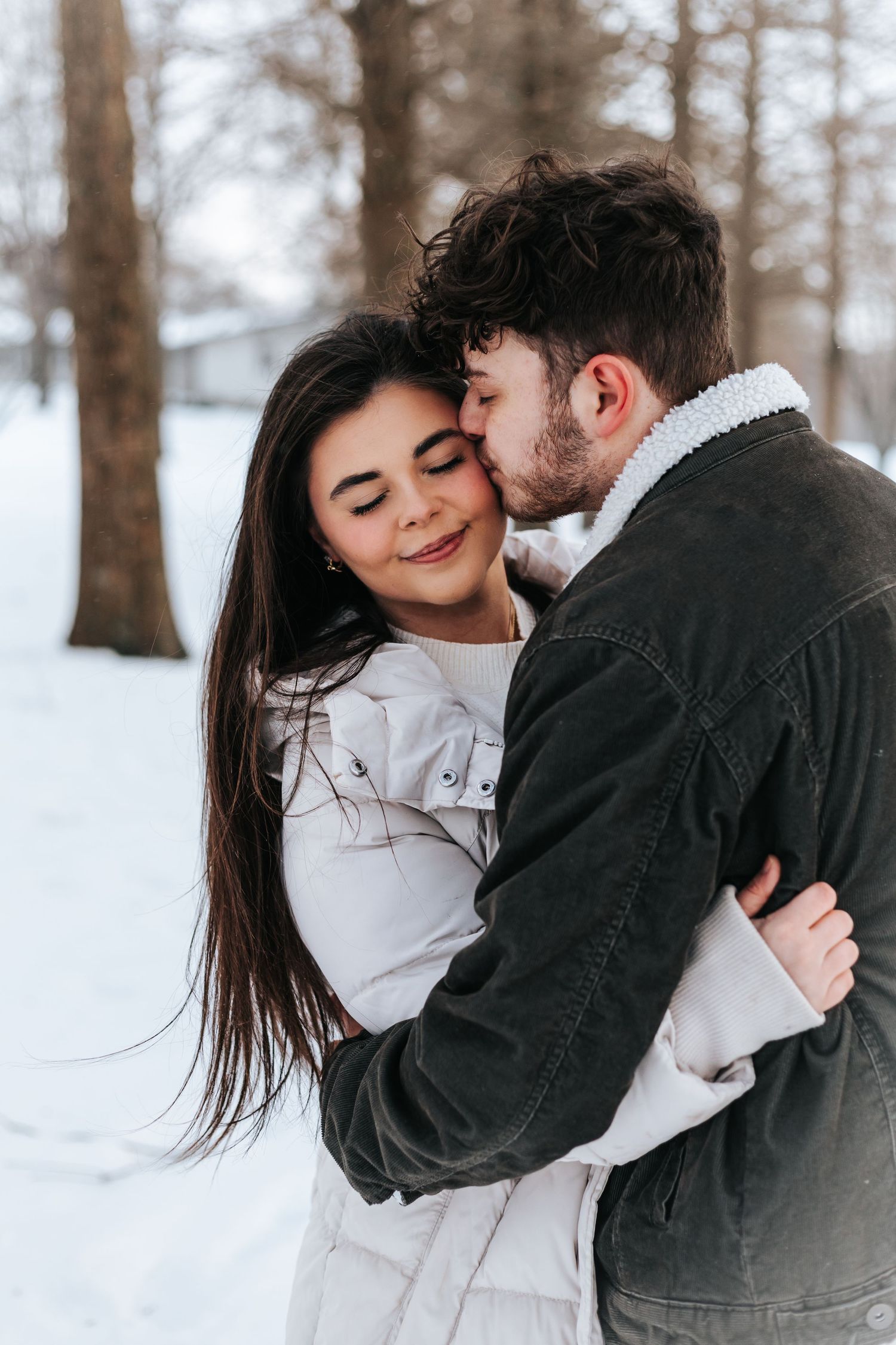 Tender winter embrace between couple wearing cozy jackets in snowy forest setting.