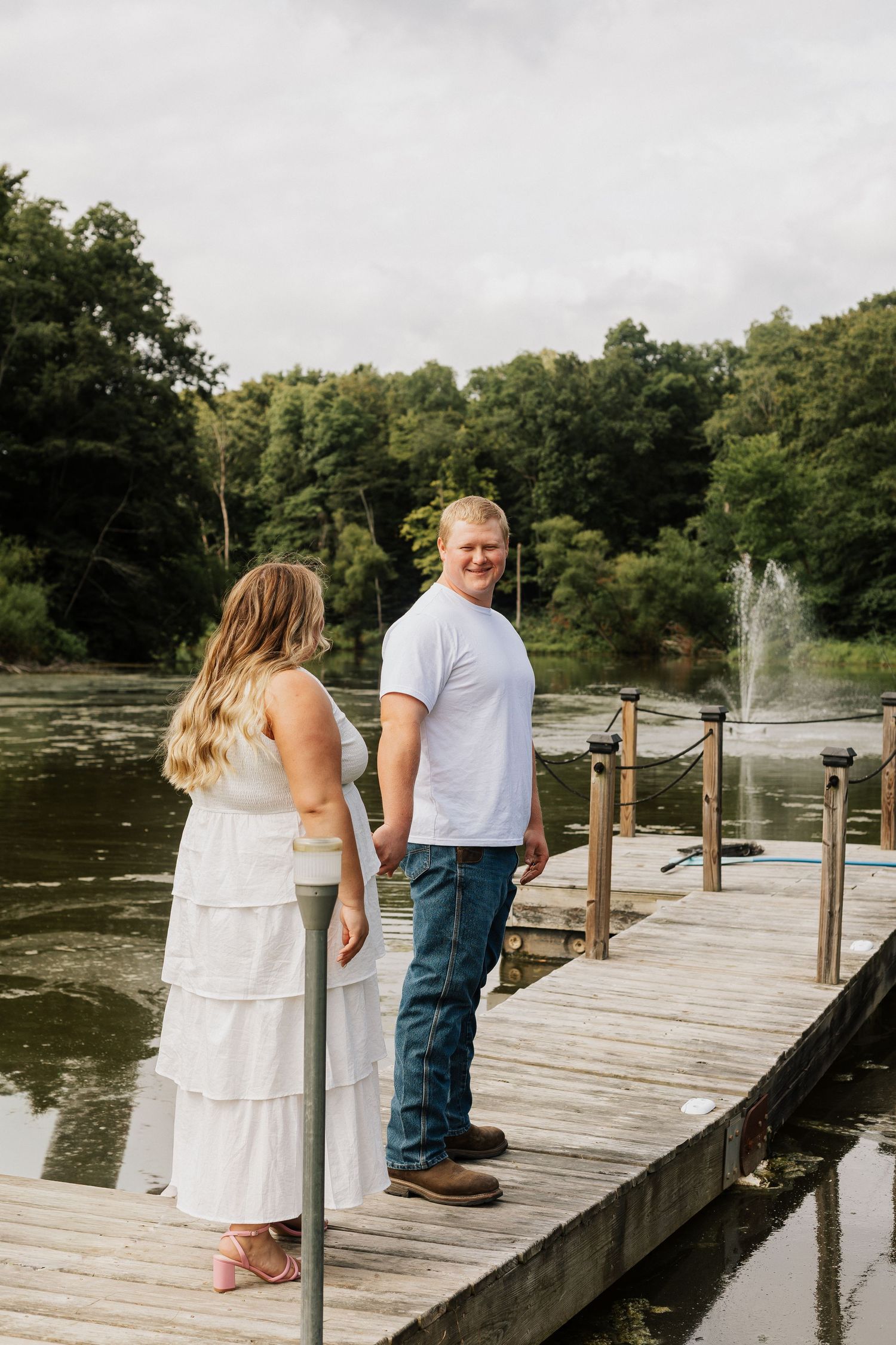 A couple stands together on a wooden dock overlooking a pond with a fountain.