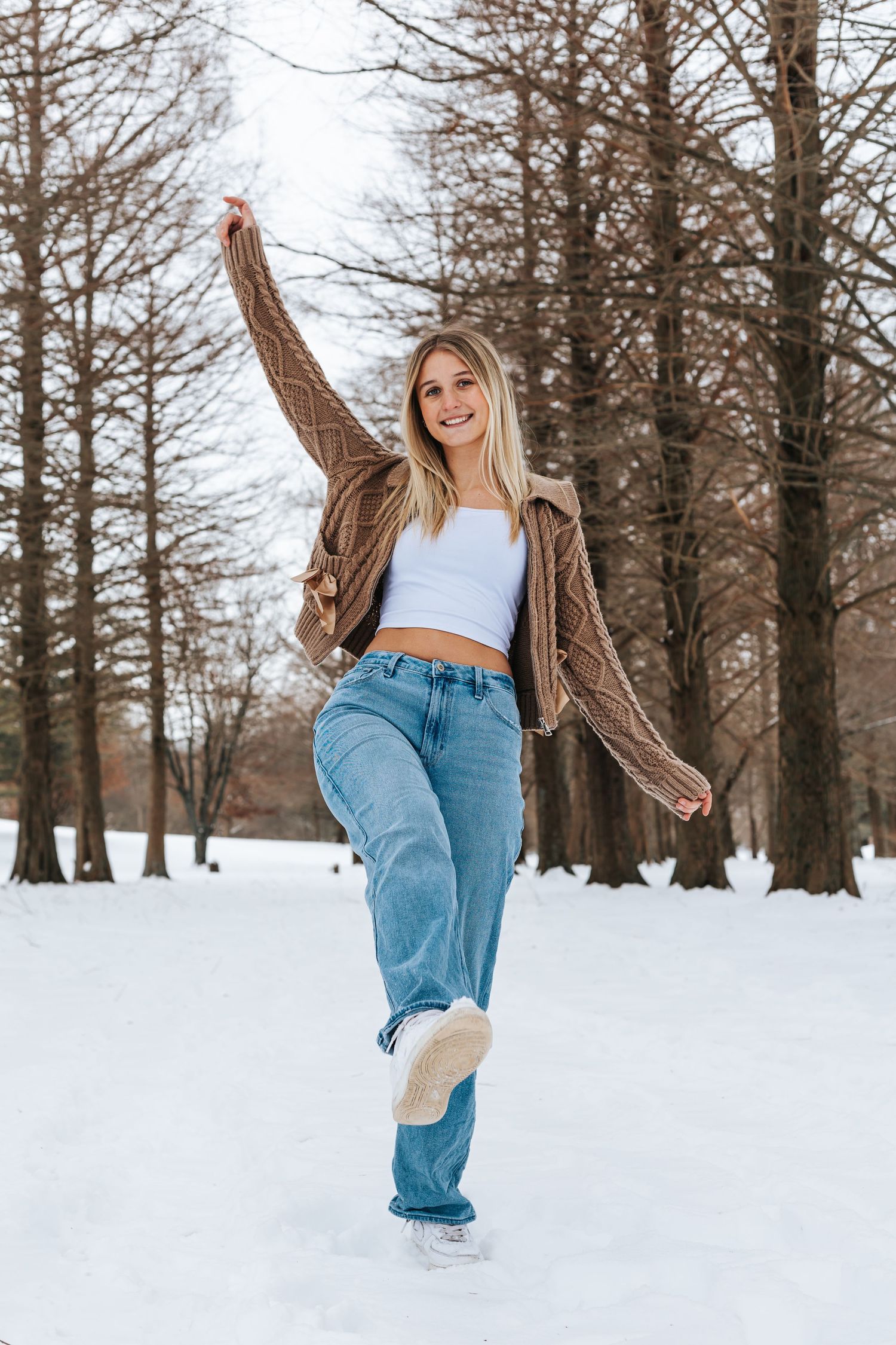 Person in casual attire playfully kicking up snow in a winter forest setting.