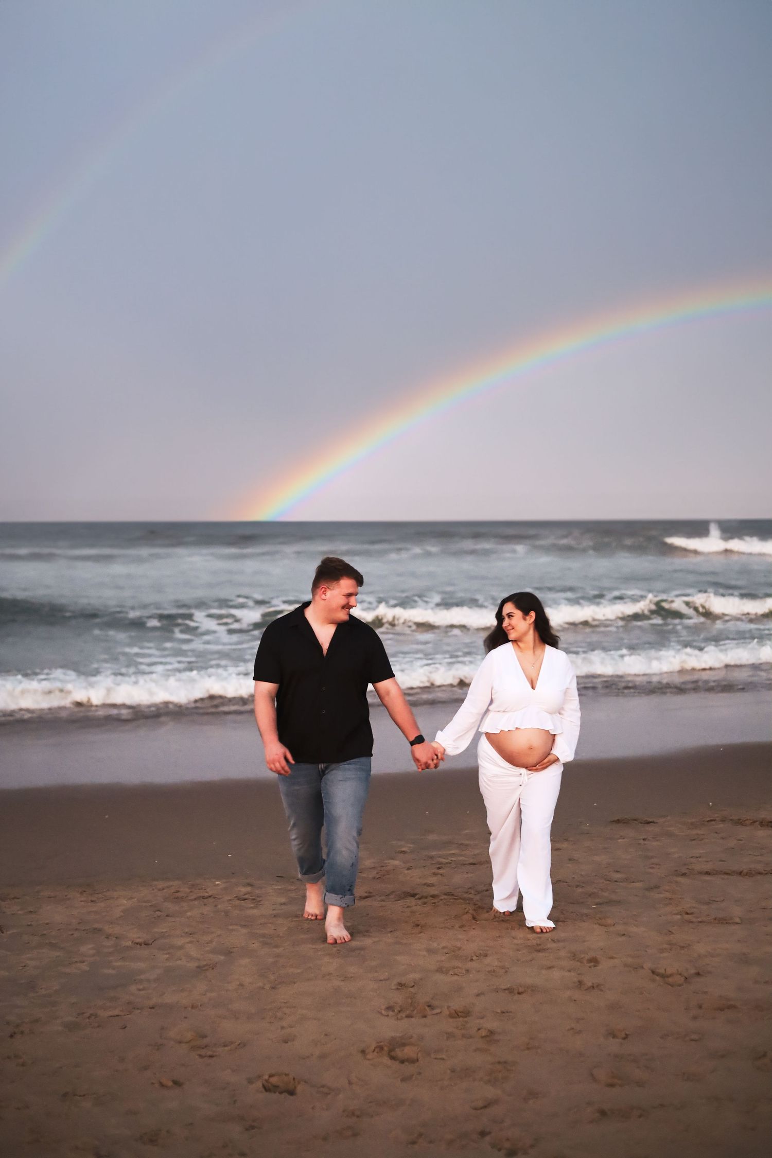 Couple walking on beach with rainbow overhead during pregnancy announcement photoshoot.