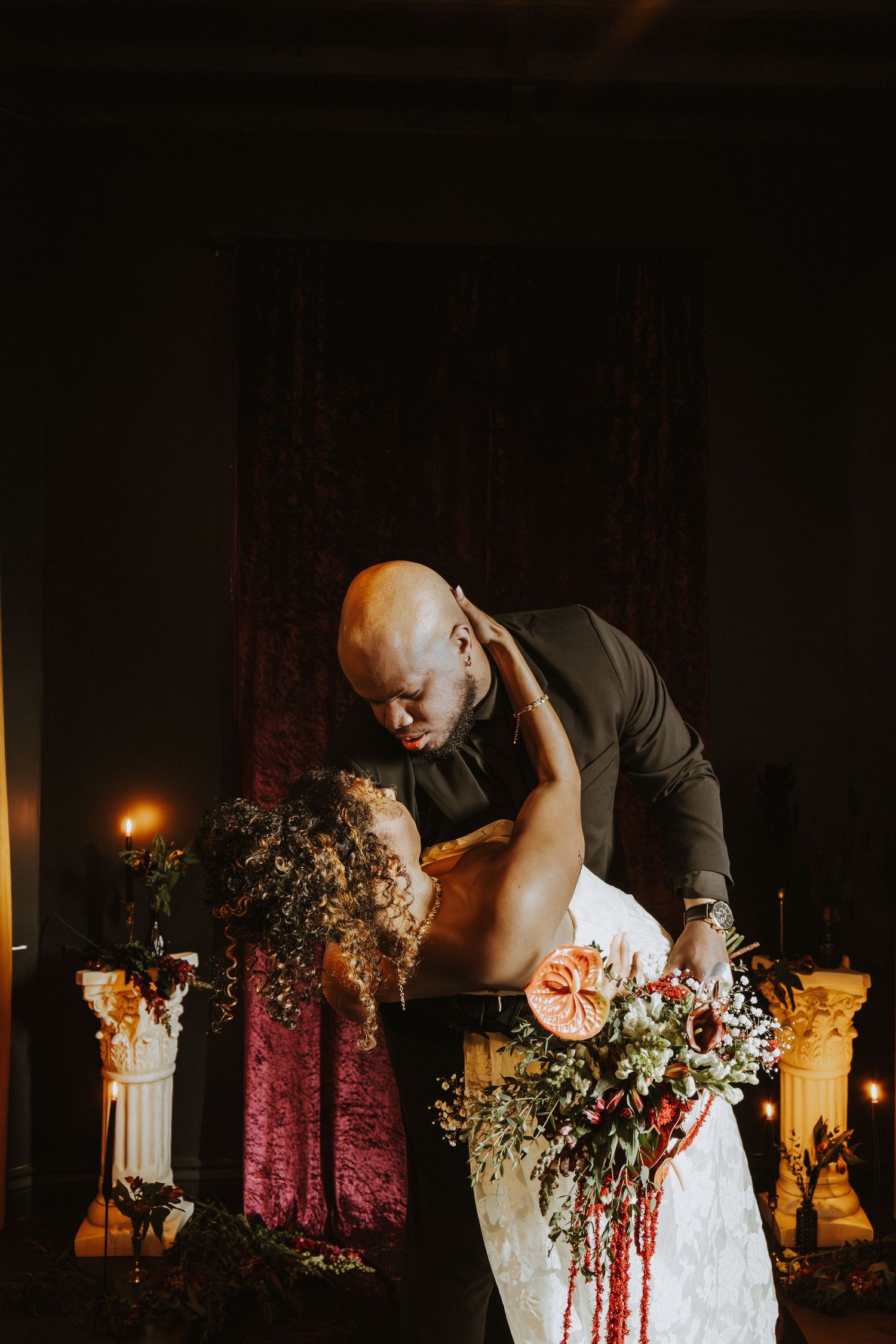 Dramatic wedding portrait with candlelit backdrop and cascading red floral bouquet.