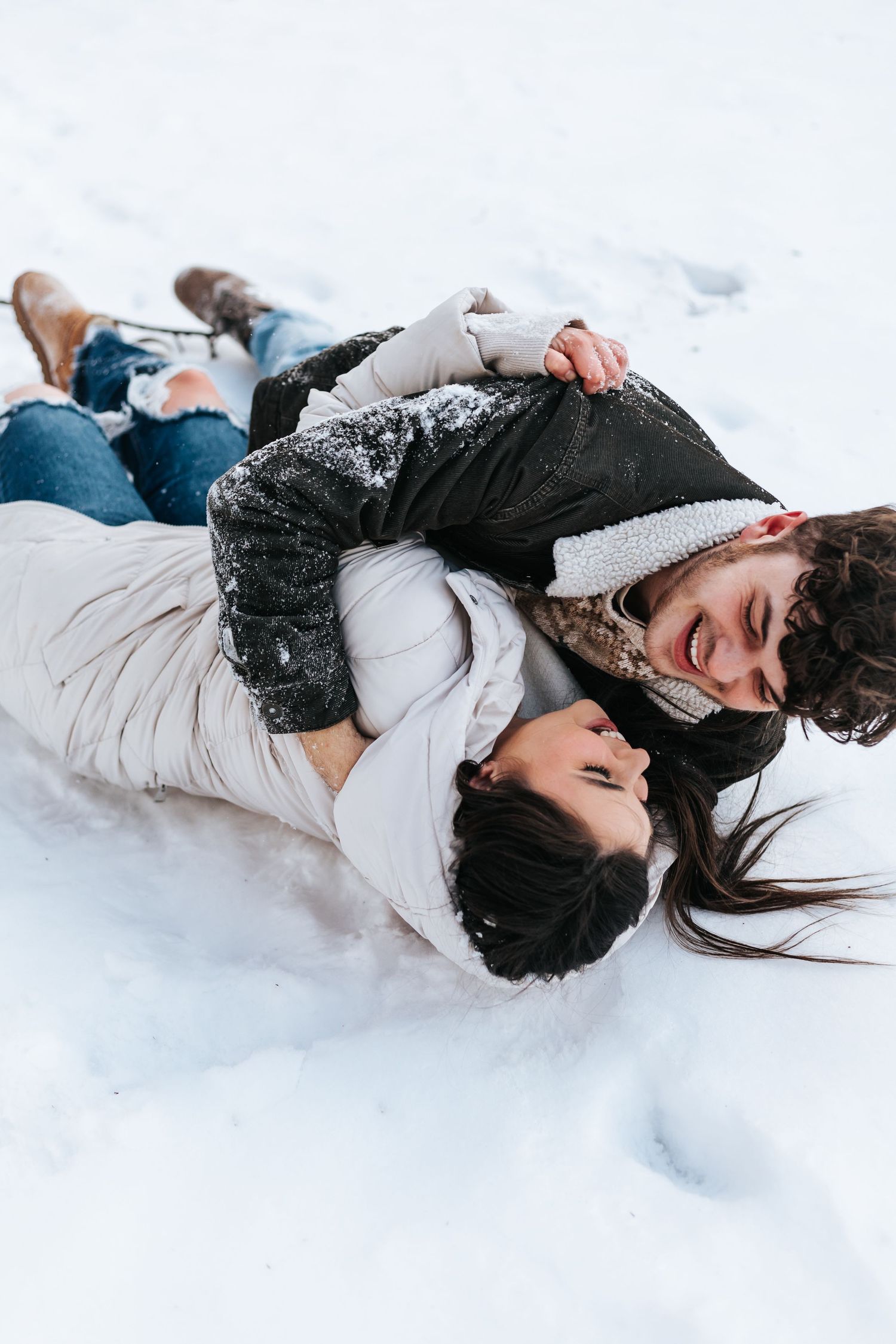 Playful couple lying in fresh snow wearing winter coats and jeans.