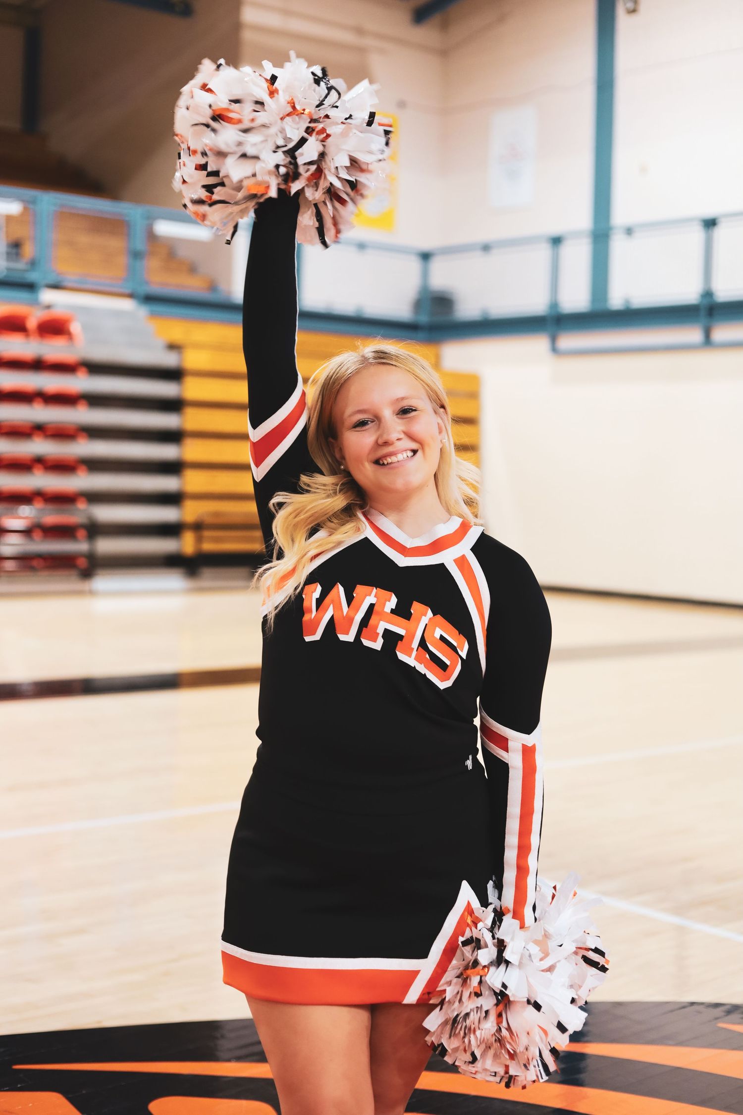 Person in cheerleading uniform poses with pom poms in gymnasium.