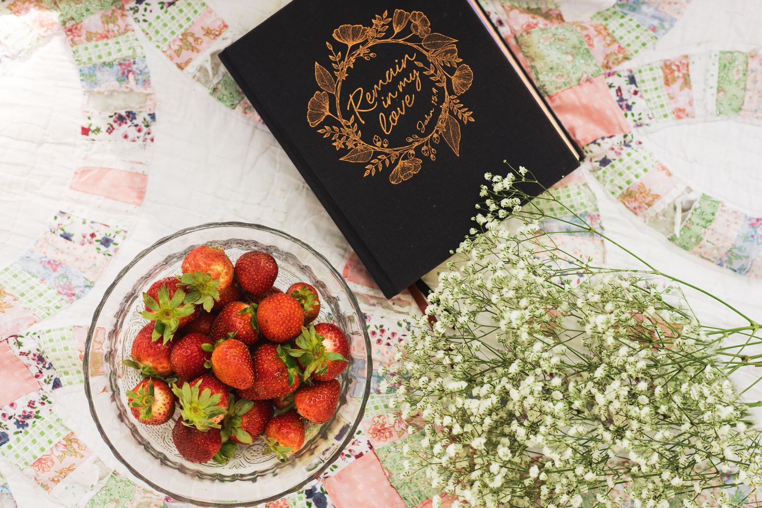 Fresh strawberries in a glass bowl next to baby's breath flowers and a black book on floral background.