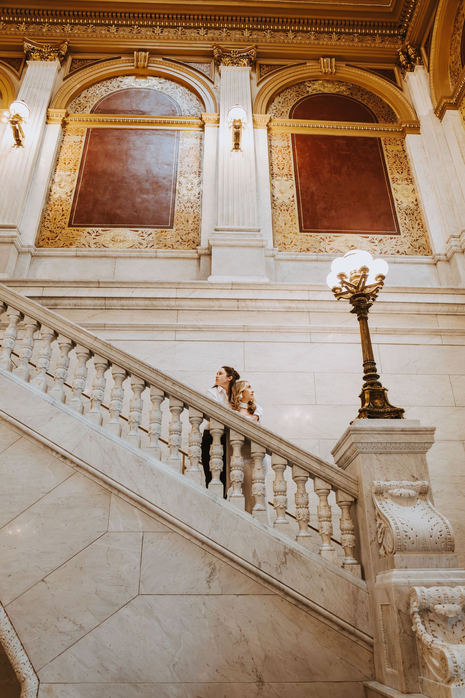 Bride walks up grand marble staircase in ornate gold and white classical building with decorative lamp post.