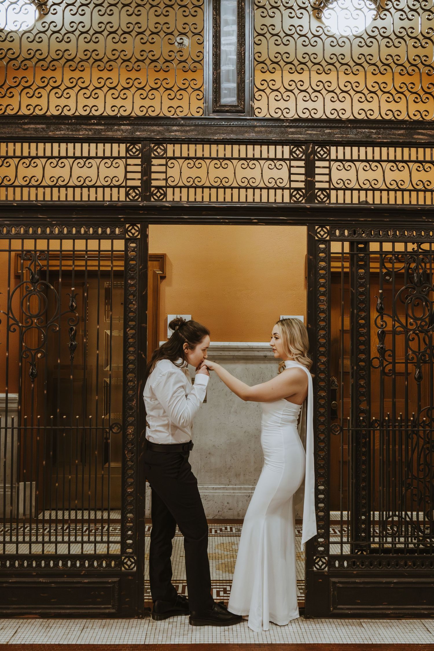 Bride in white gown adjusts groom suit at ornate iron gate doorway in elegant wedding portrait.