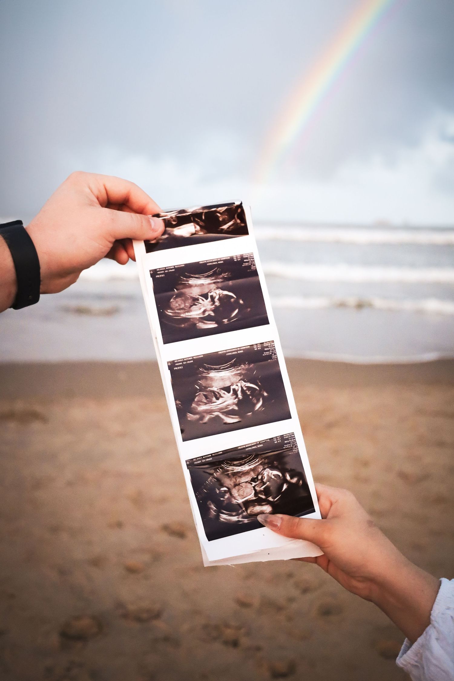 Hands holding ultrasound photos on beach with rainbow in background.