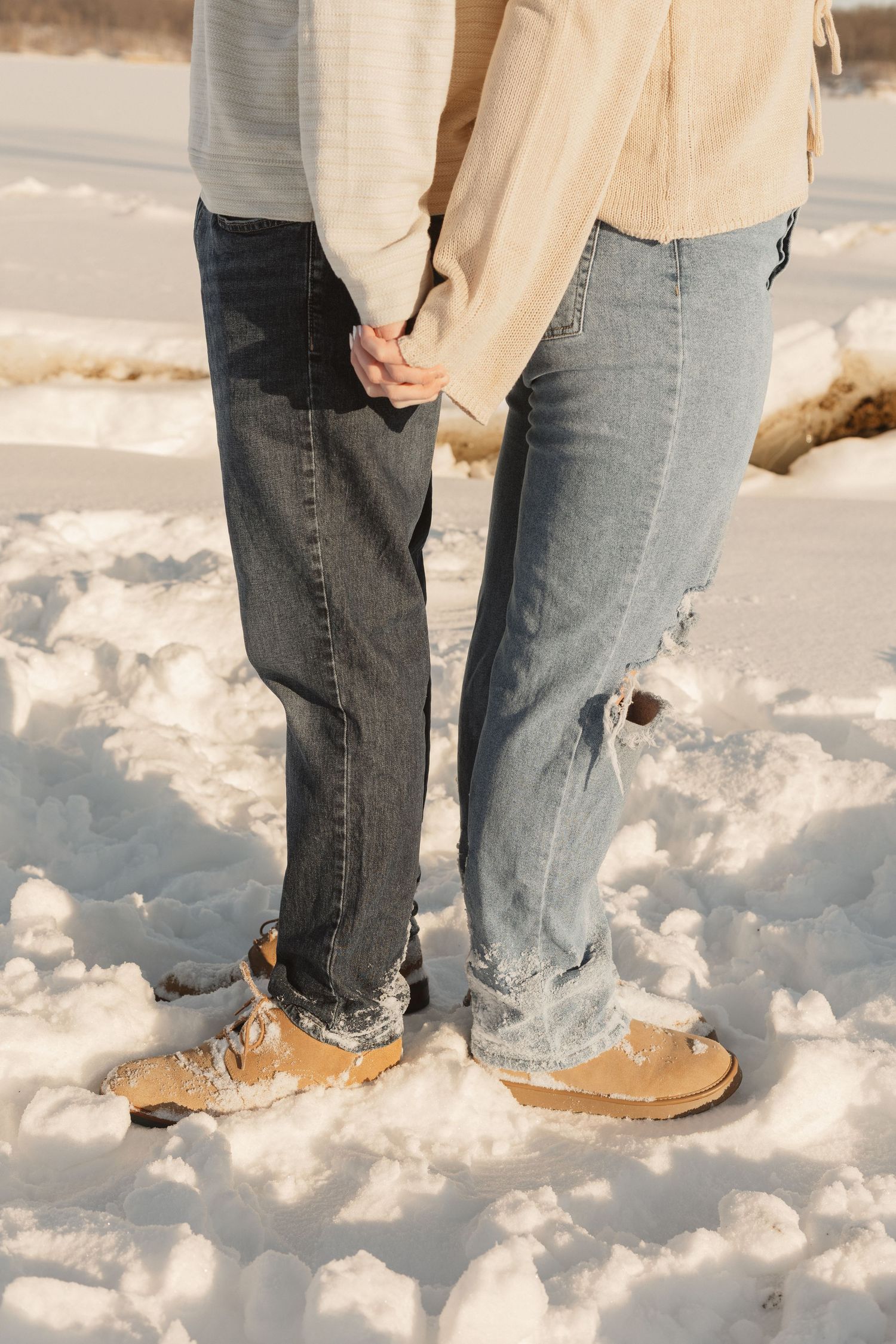 Two pairs of boots stand in fresh snow wearing jeans and tan jackets on a winter day.