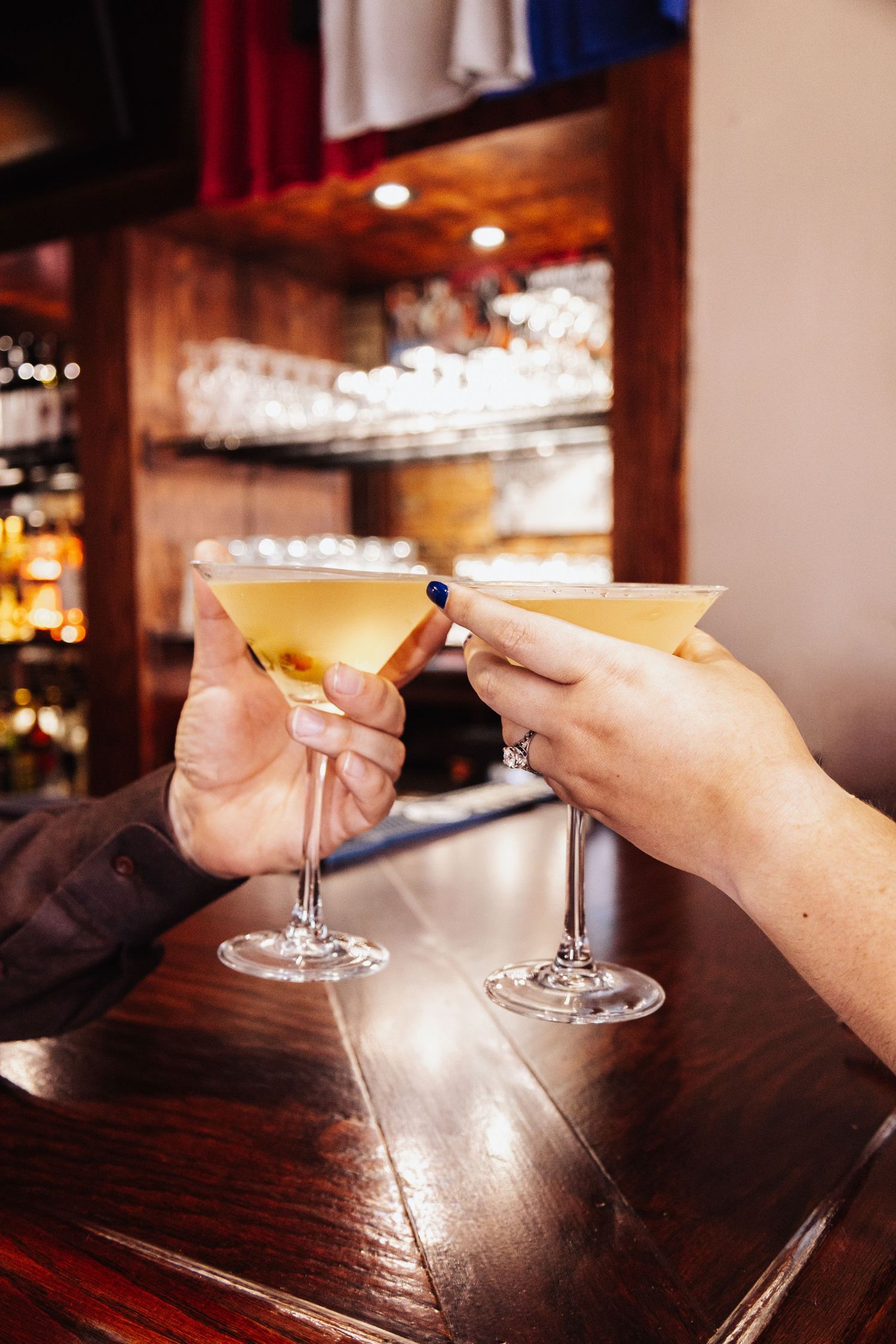 Martini glasses clink together in a toast at a dimly lit bar.