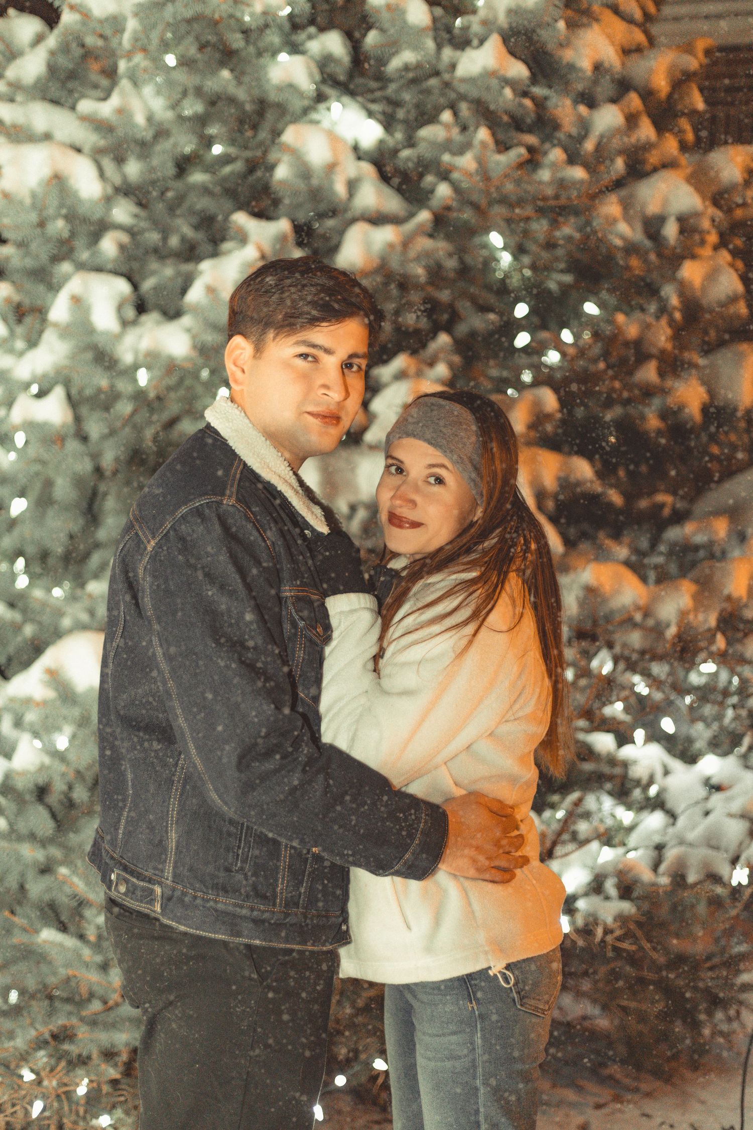 A couple sharing an intimate moment in front of snow-covered evergreen trees at night.