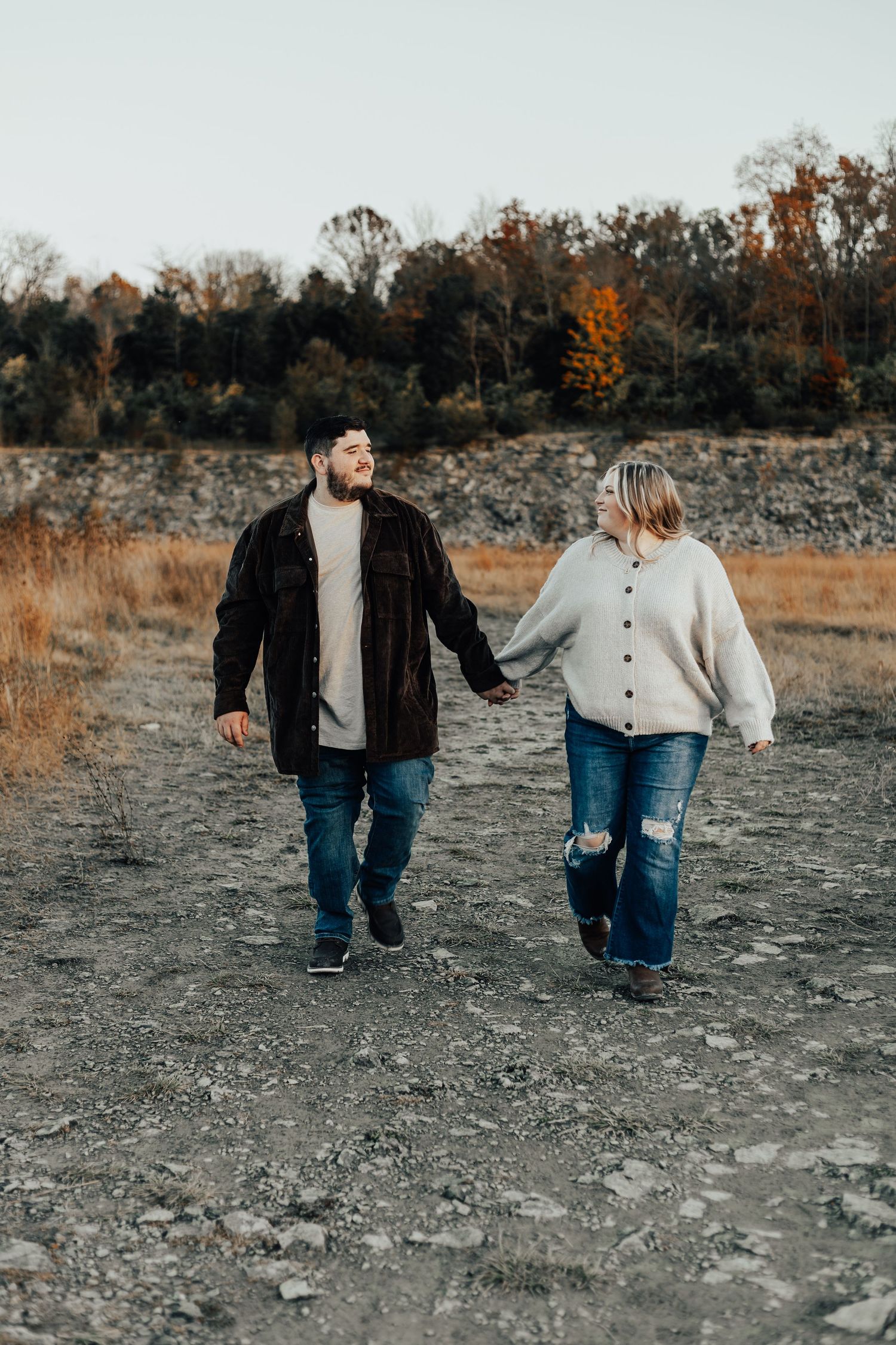 Couple holding hands while walking on rocky terrain with autumn trees in background.