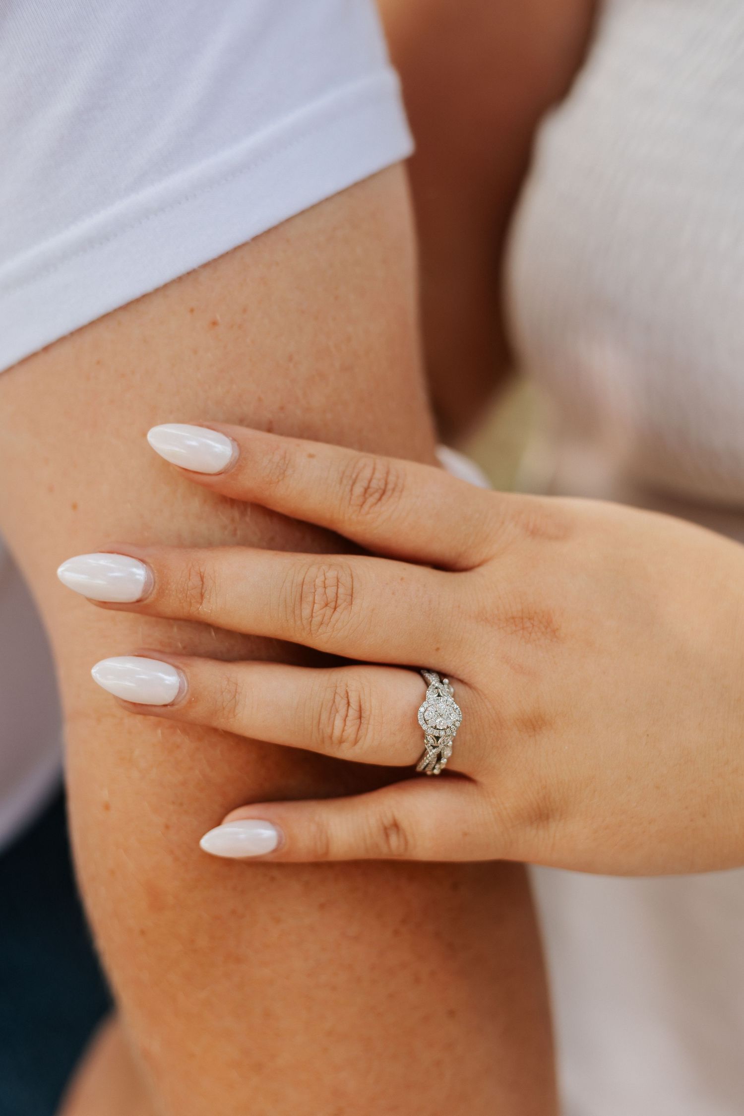A hand wearing white nail polish displays a sparkling engagement ring.
