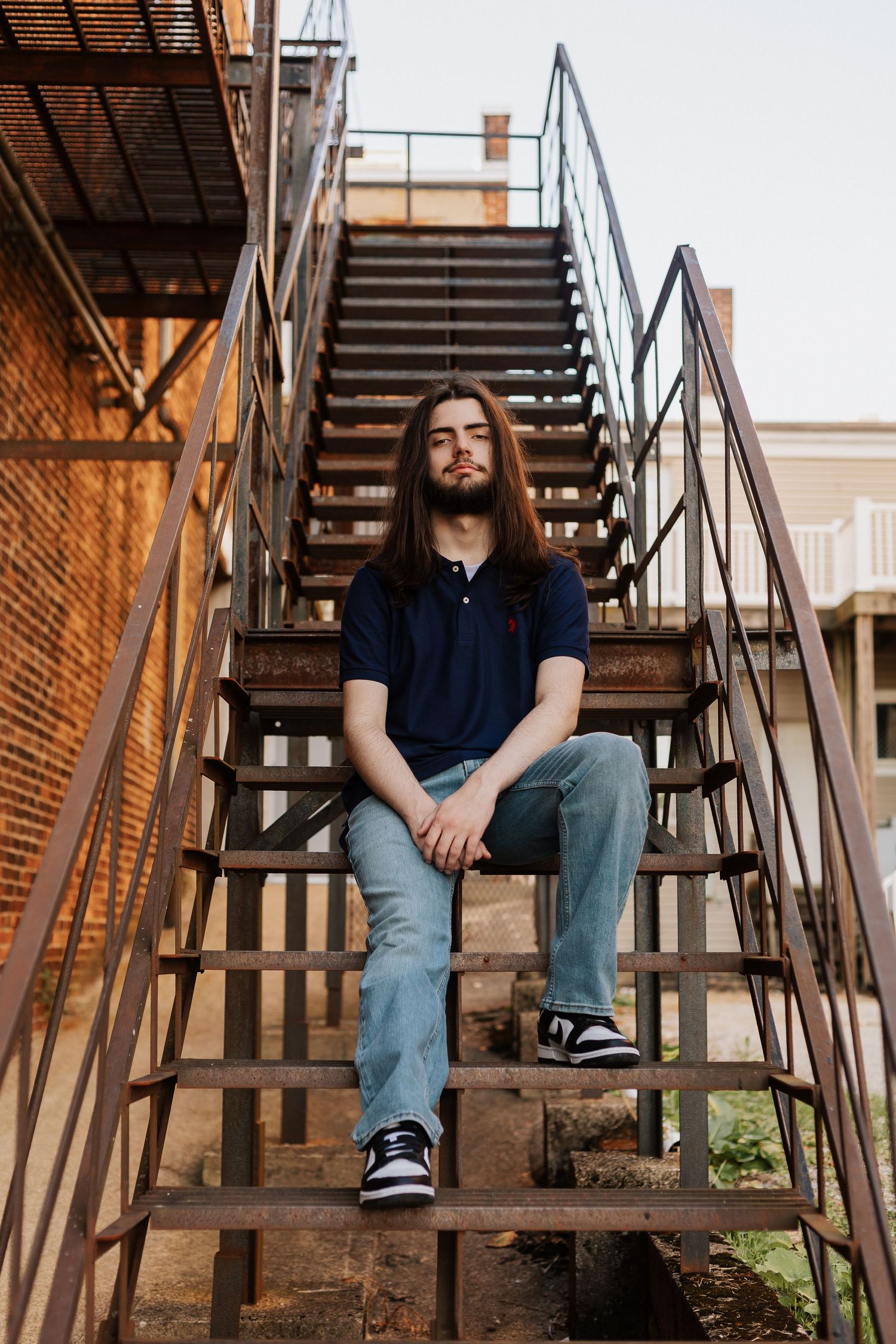 Person sitting on rustic metal fire escape stairs against brick wall in casual attire.