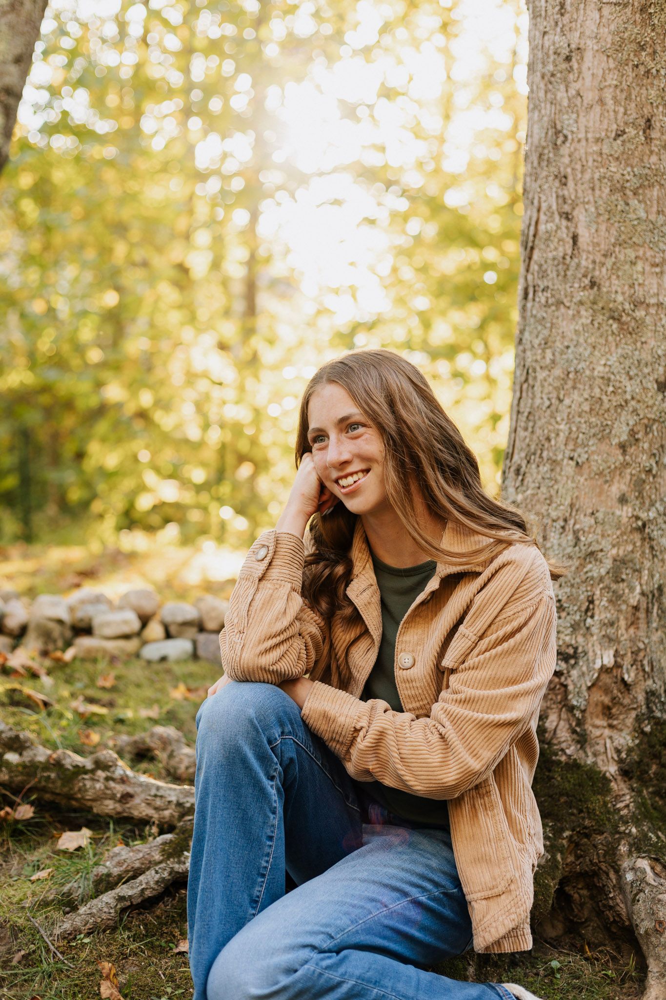 A person in a tan jacket and jeans sits against a tree trunk in a sunlit autumn forest setting.