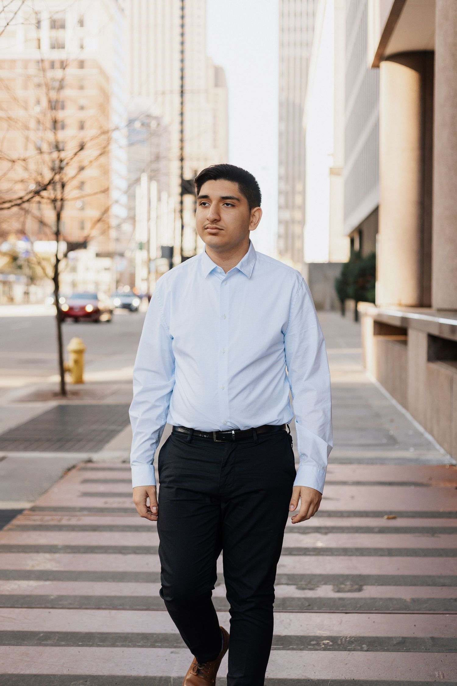 Professional business portrait of person in light blue dress shirt and dark pants standing in urban setting.