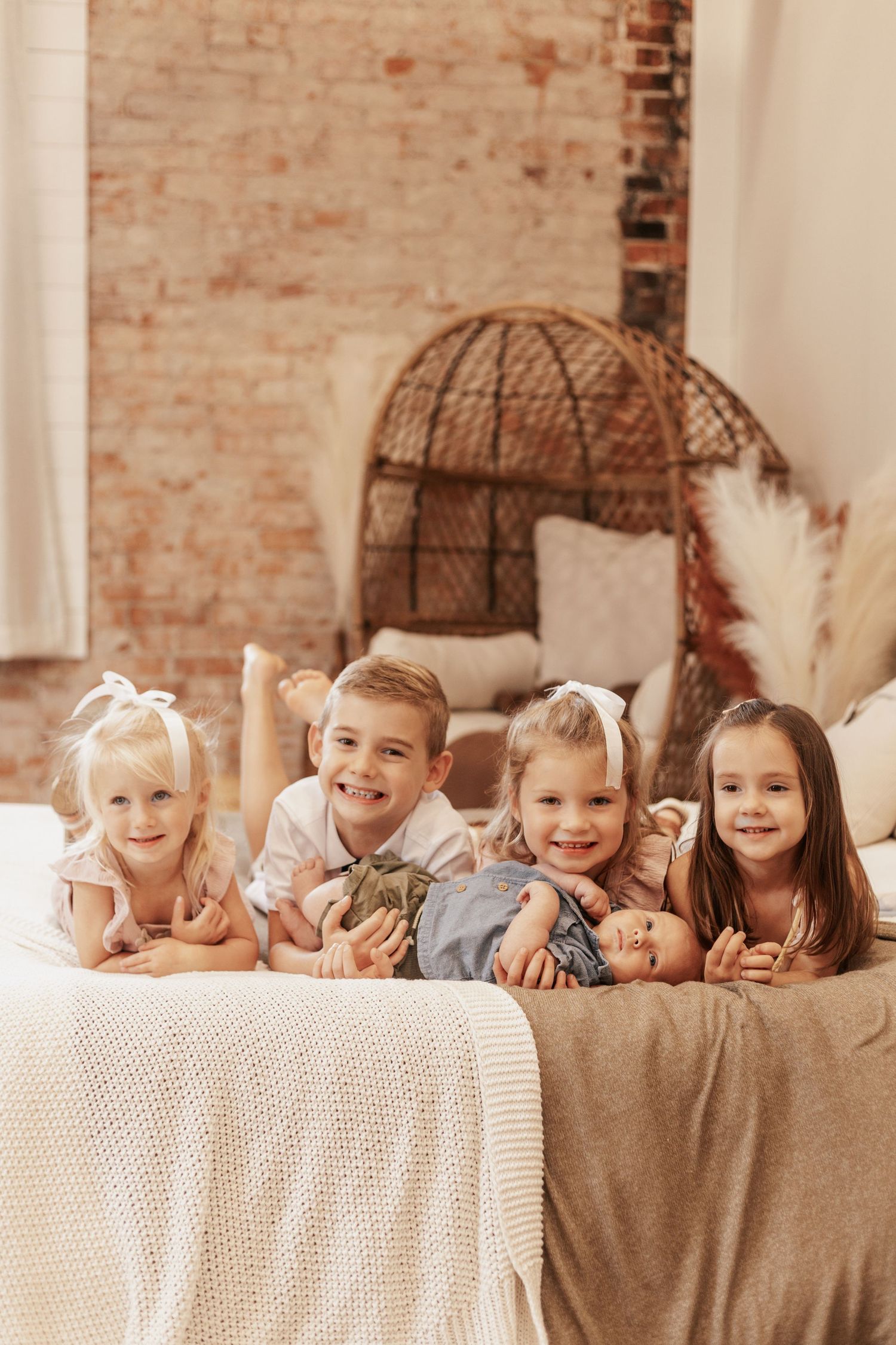 Group of children lying on bed with wicker chair and exposed brick wall behind them.