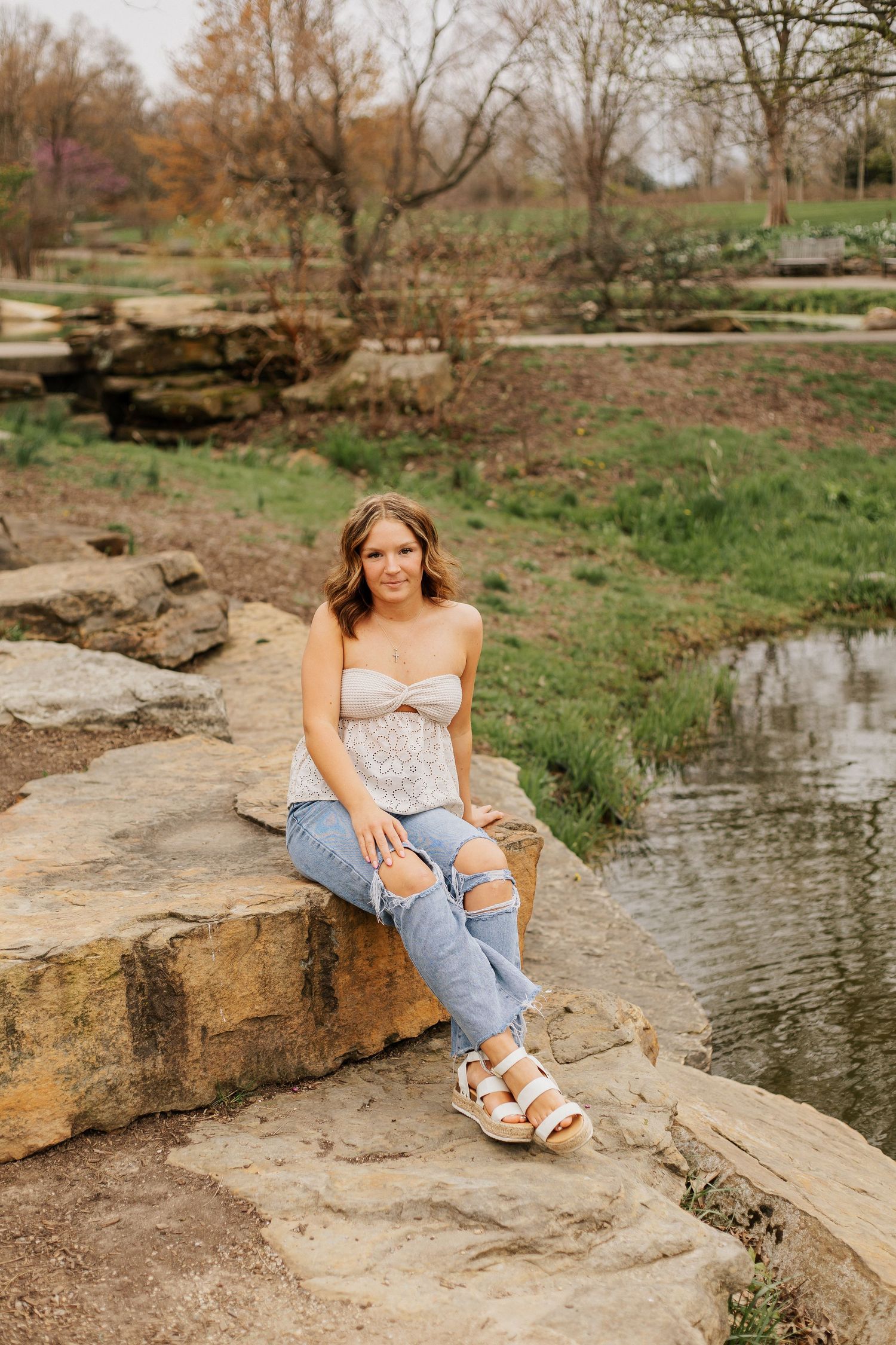 A person in jeans and white top poses casually on large rocks beside a peaceful pond on a spring day.