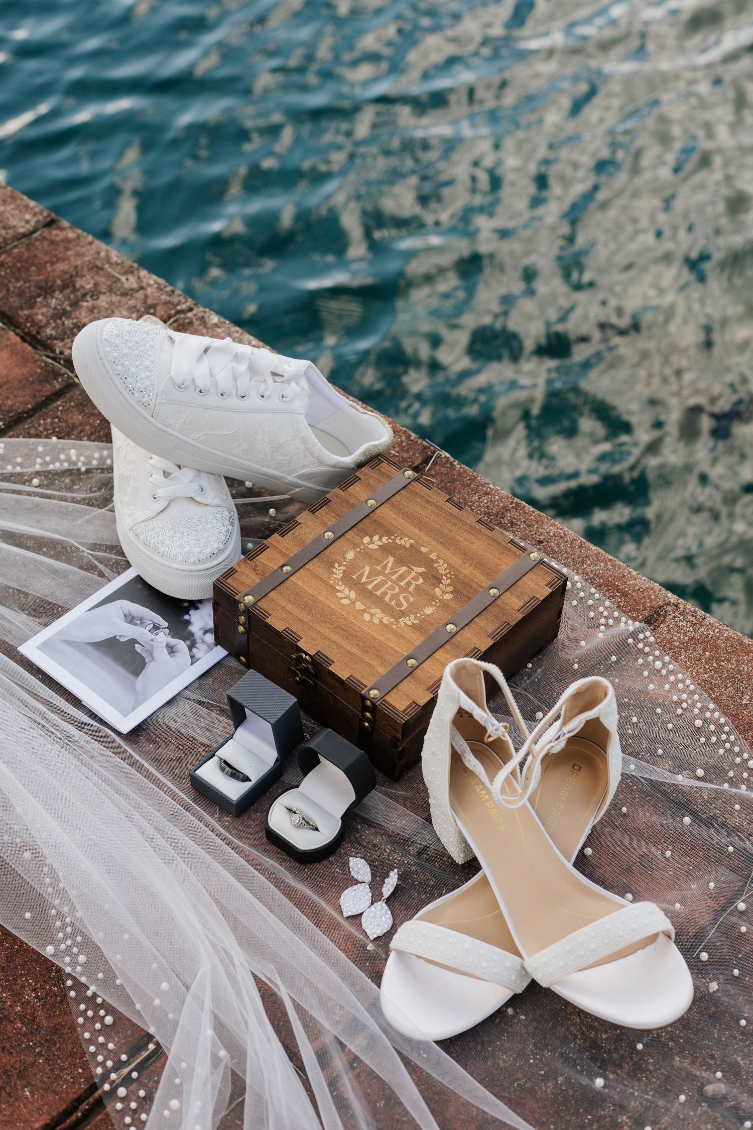 Wedding accessories including white shoes, veil, and wooden box arranged on a stone surface near water.