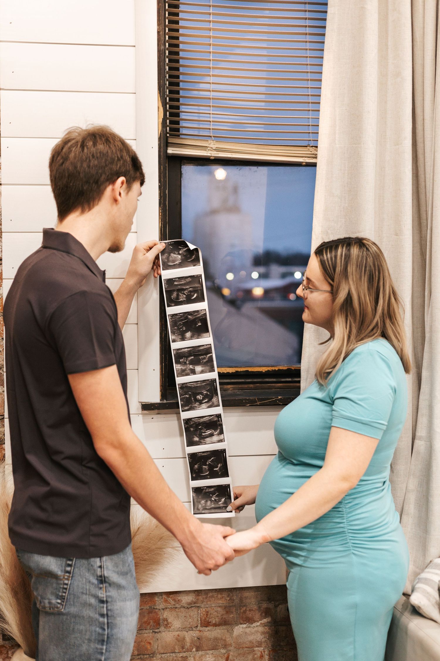 Expectant couple looking at ultrasound photos together against a brick wall.