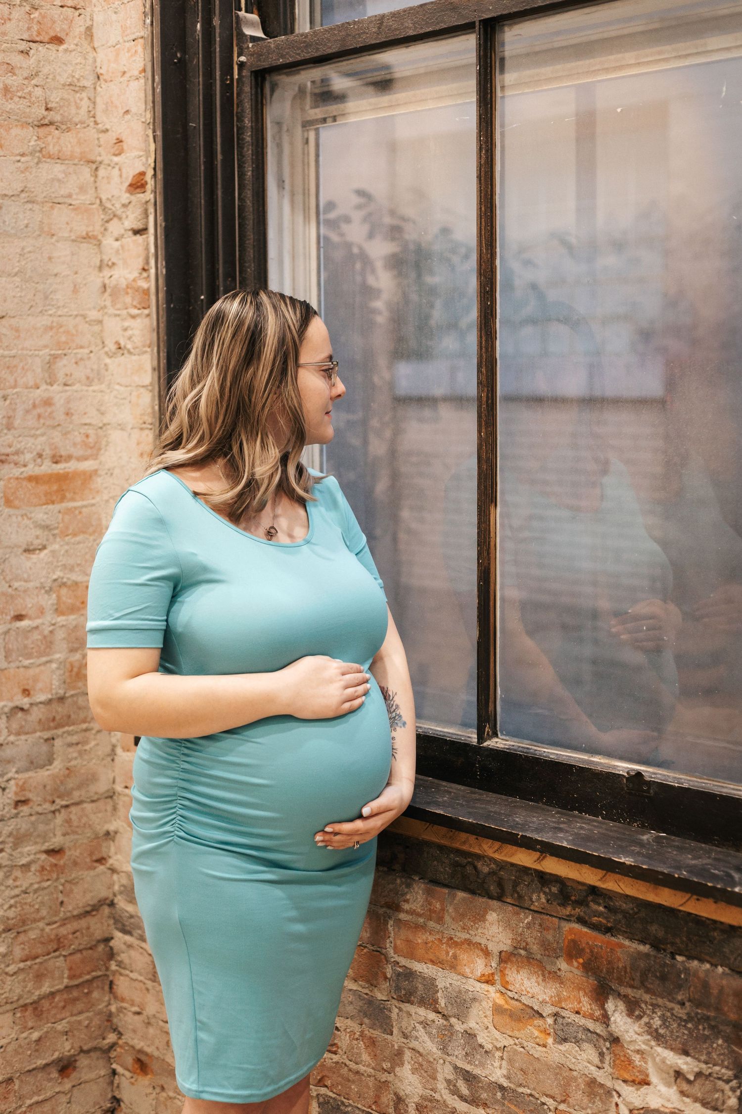Person in turquoise dress standing by industrial windows with exposed brick wall.