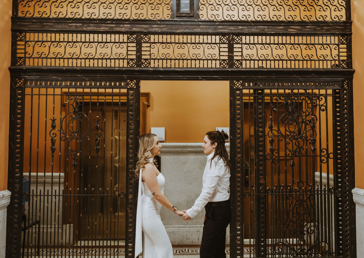 Two brides hold hands before ornate iron gates in a romantic wedding portrait.