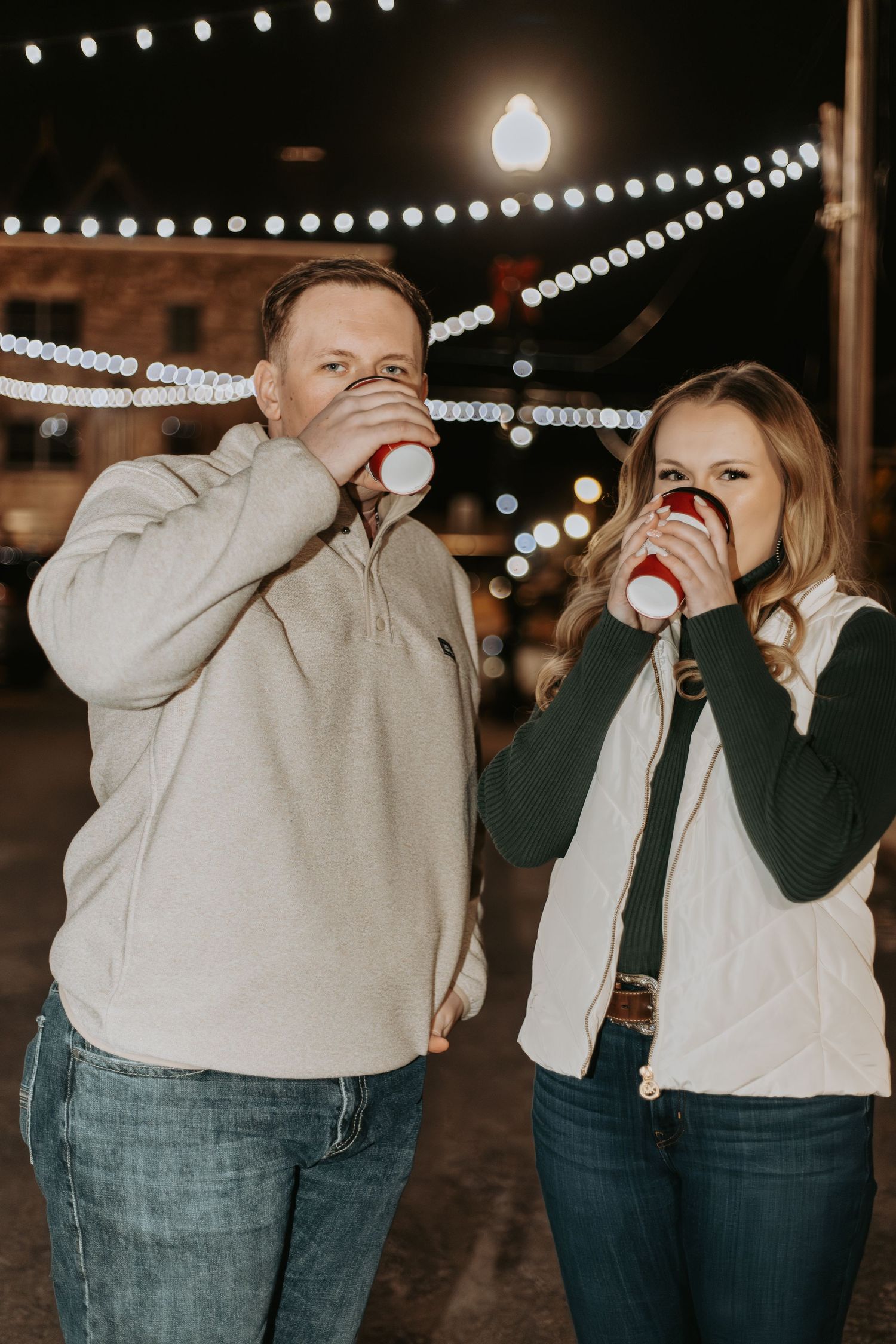 Playful couple drinking from coffee cups under string lights at night.