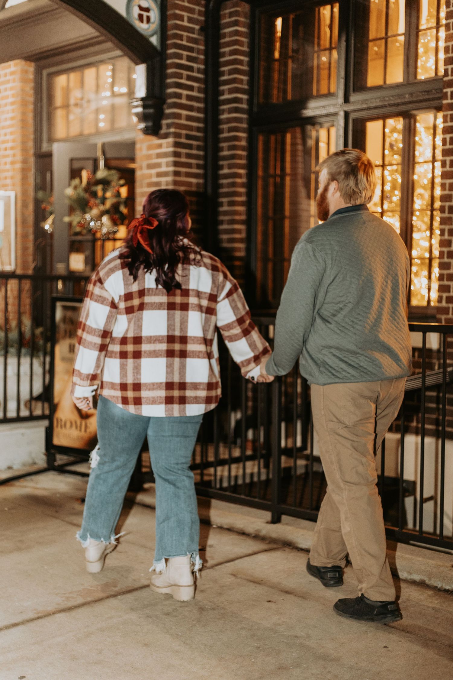 A couple walks hand in hand along an illuminated storefront at night.