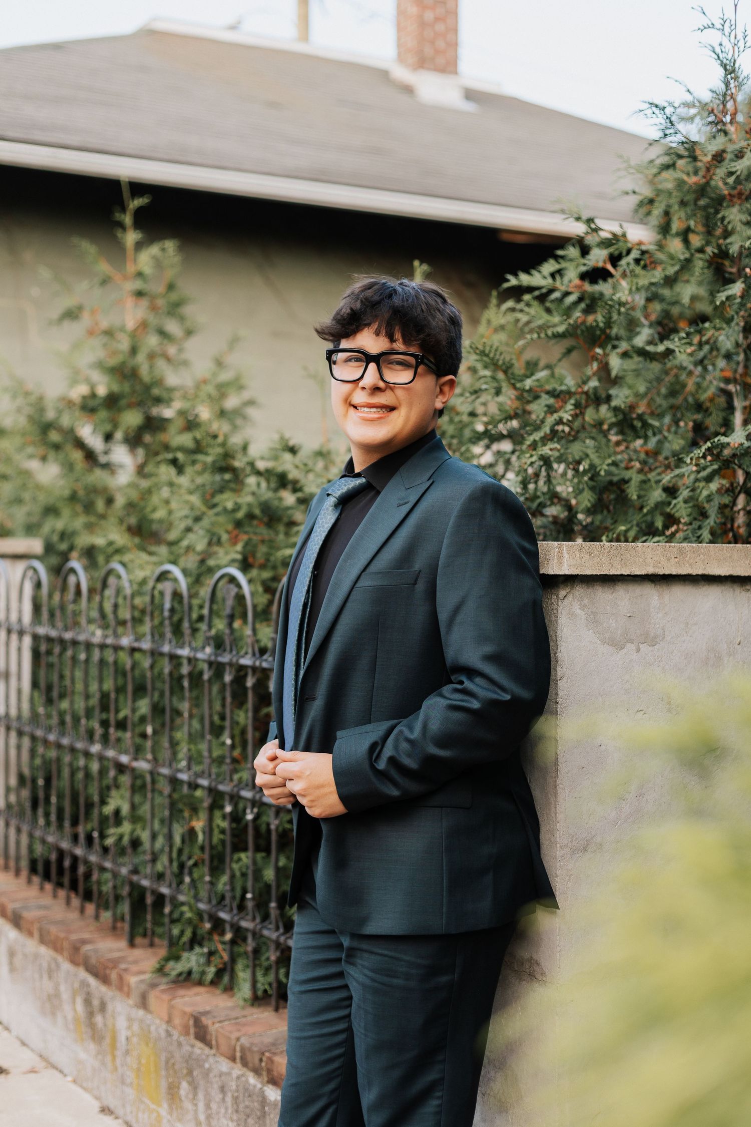 Professional portrait of a person in dark suit and glasses standing by garden fence.